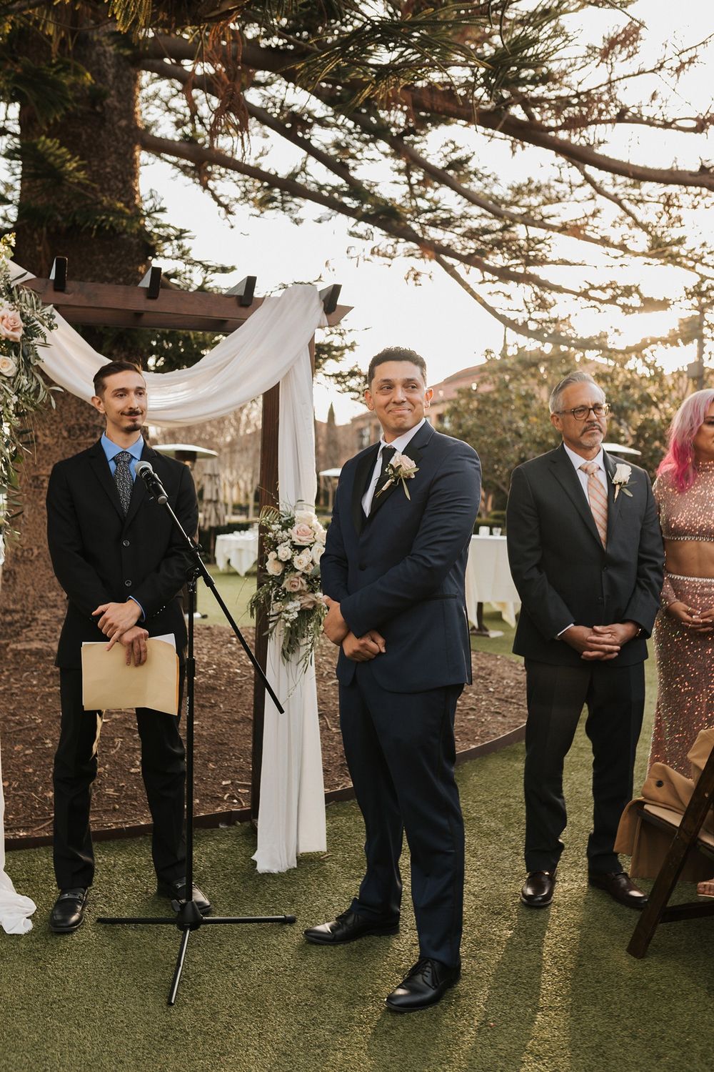 A groom waits for his bride at an outdoor wedding ceremony under a pine tree at Stone Brewing Liberty Station in San Diego.