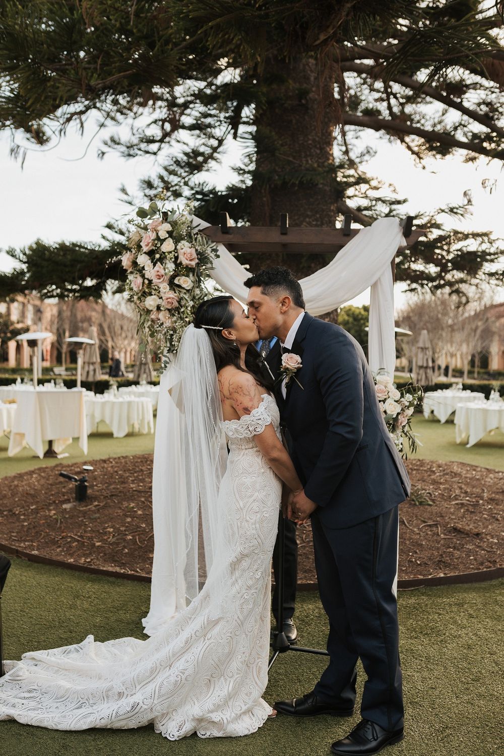 A couple share their first kiss at a romantic outdoor wedding ceremony under a pine tree at Stone Brewing Liberty Station in San Diego.