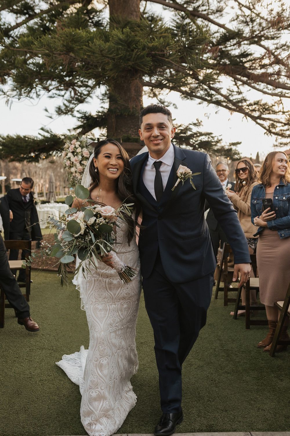 A joyful couple walks together on a grassy lawn during their outdoor wedding celebration with guests in the background at Stone Brewing.