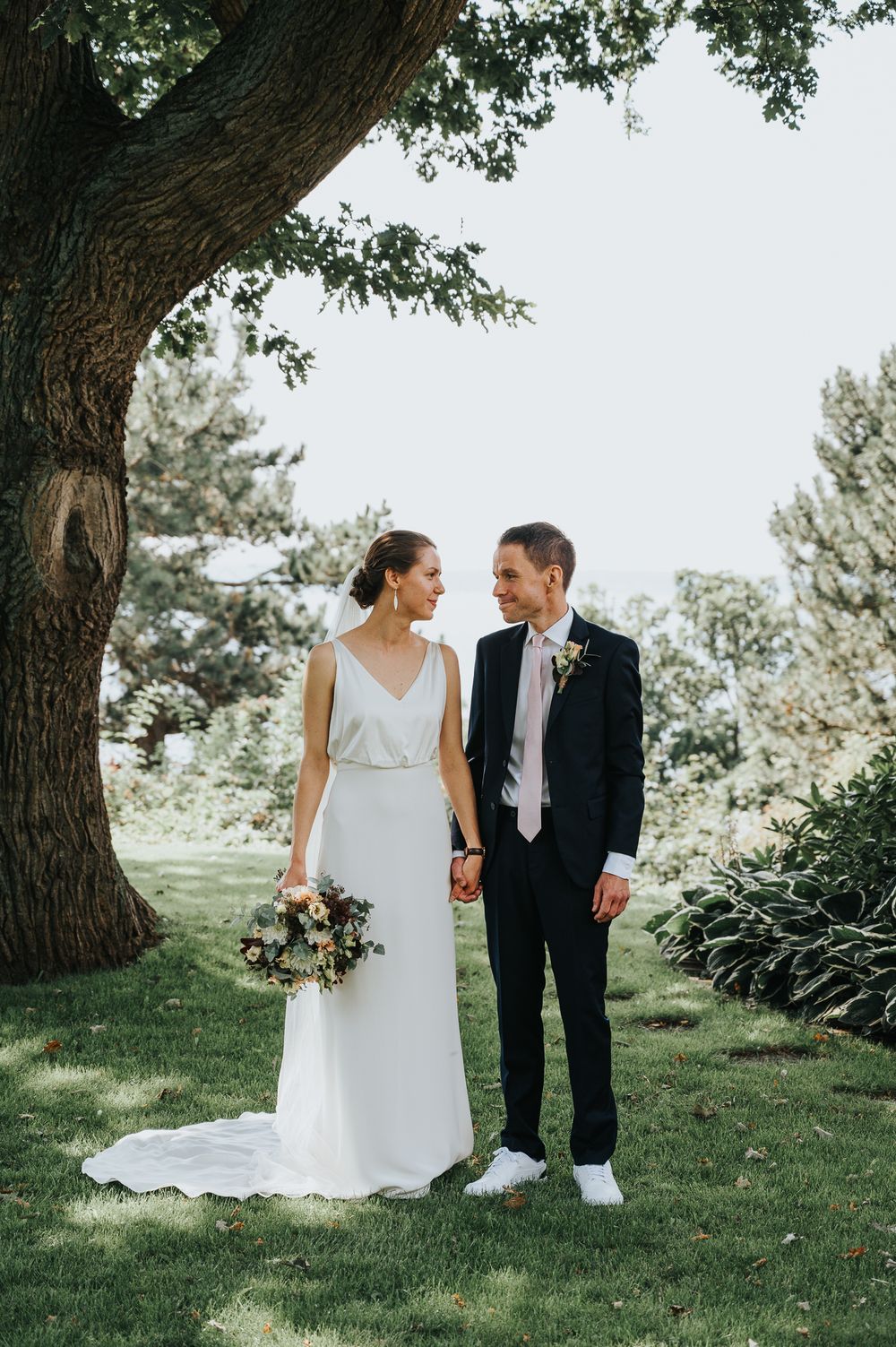 A couple shares a joyful moment under a large tree on their wedding day, with the bride holding a beautiful bouquet.