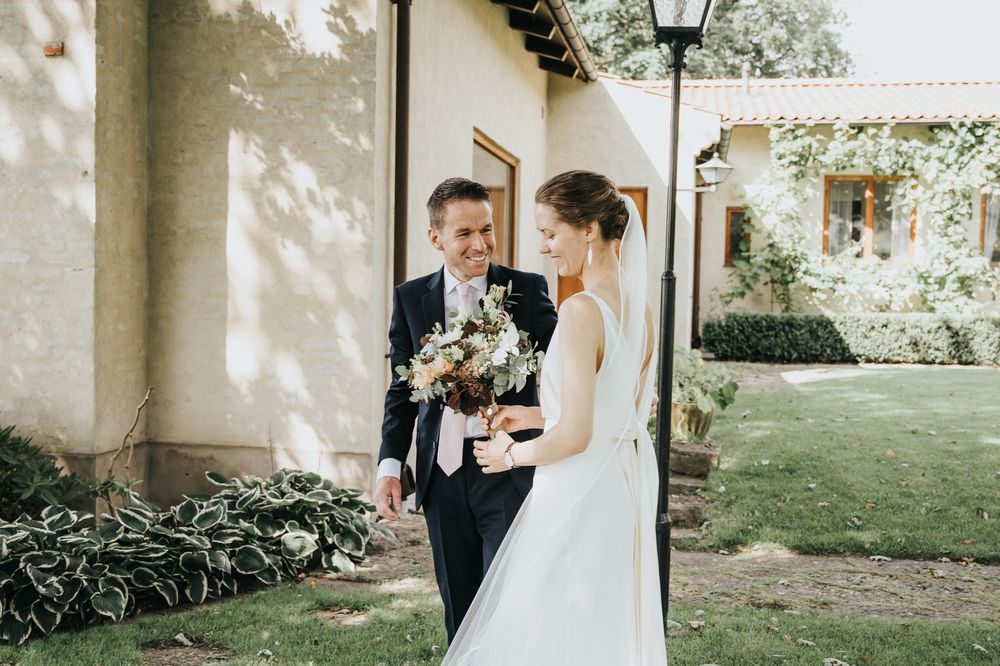 A wedding couple stands together in a garden setting holding a floral bouquet.
