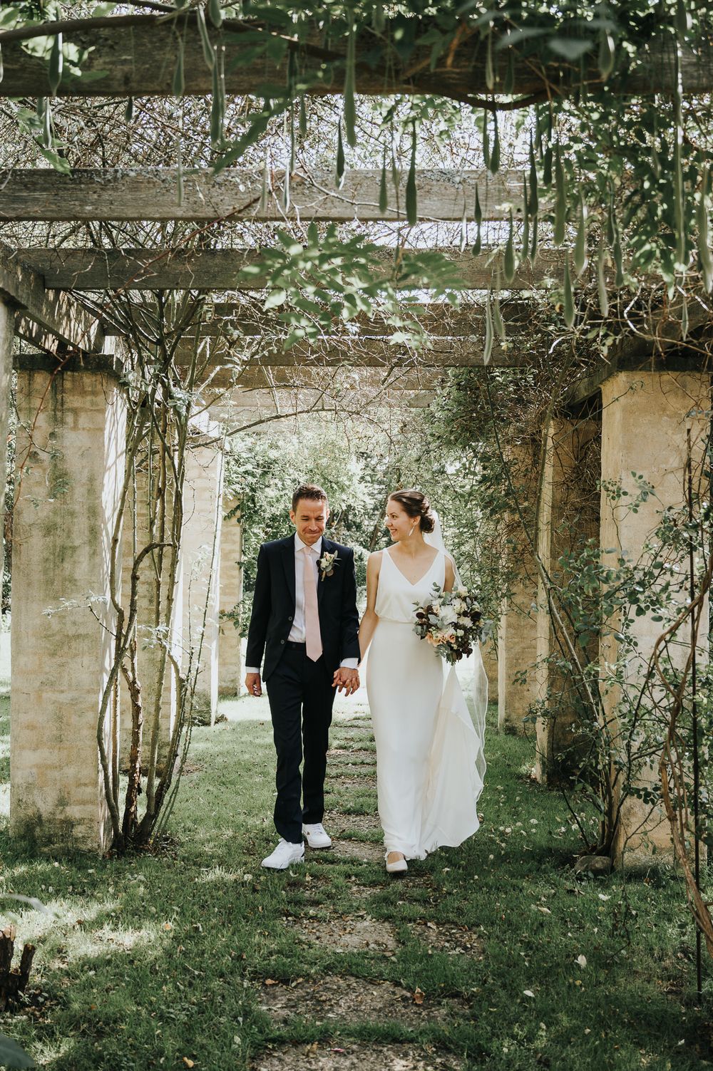 A couple walks through a romantic garden pergola covered in climbing vines and greenery.