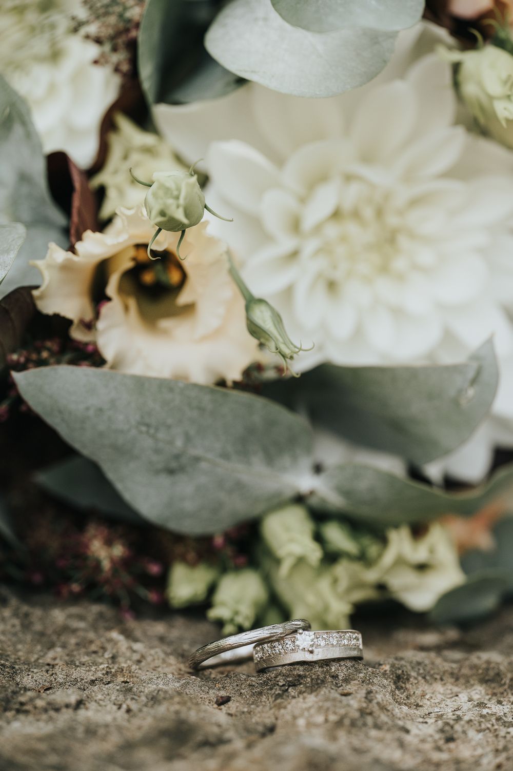 Close-up macro shot of a diamond wedding ring resting on stone with white flowers in soft focus background.