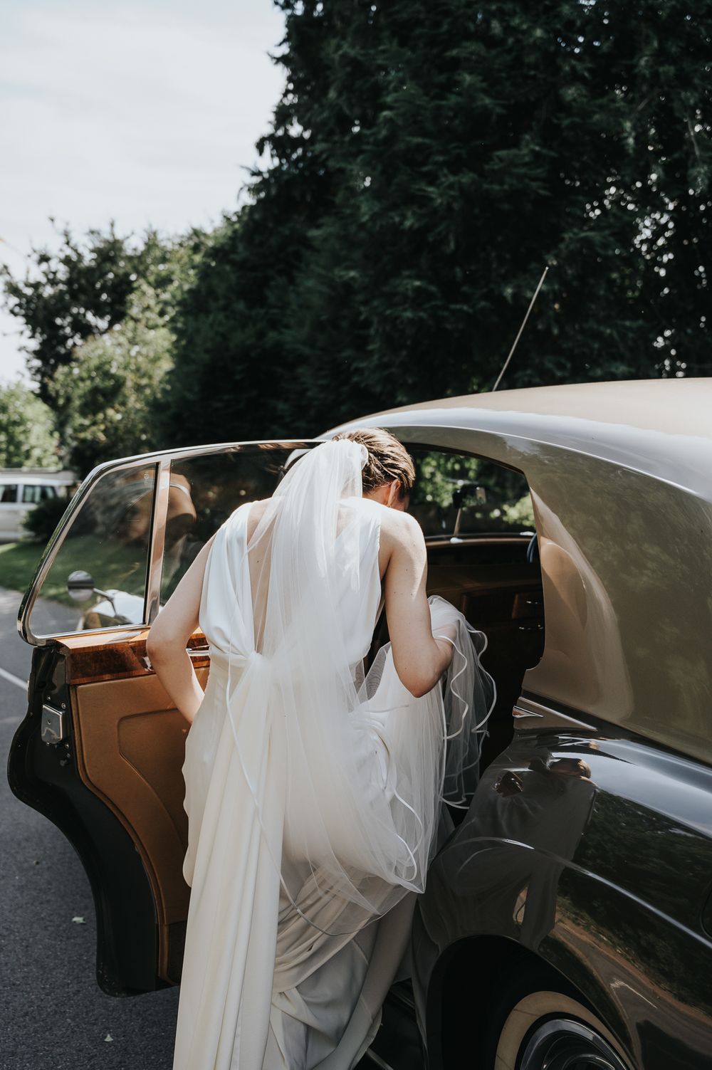 A bride in a flowing white dress and veil enters a classic vintage car with tan leather interior.
