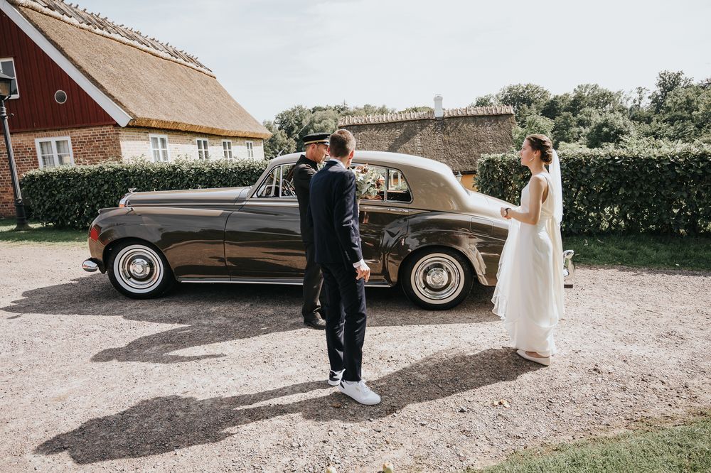 A classic vintage brown car is parked outside a red barn building during a wedding photoshoot.