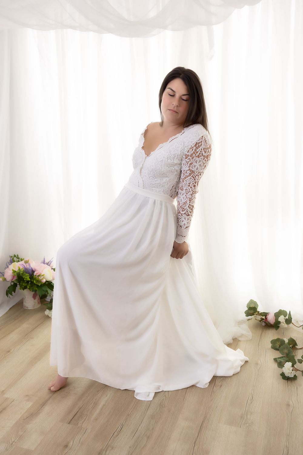A woman in a long white lace dress standing in a bright room with floral decor.