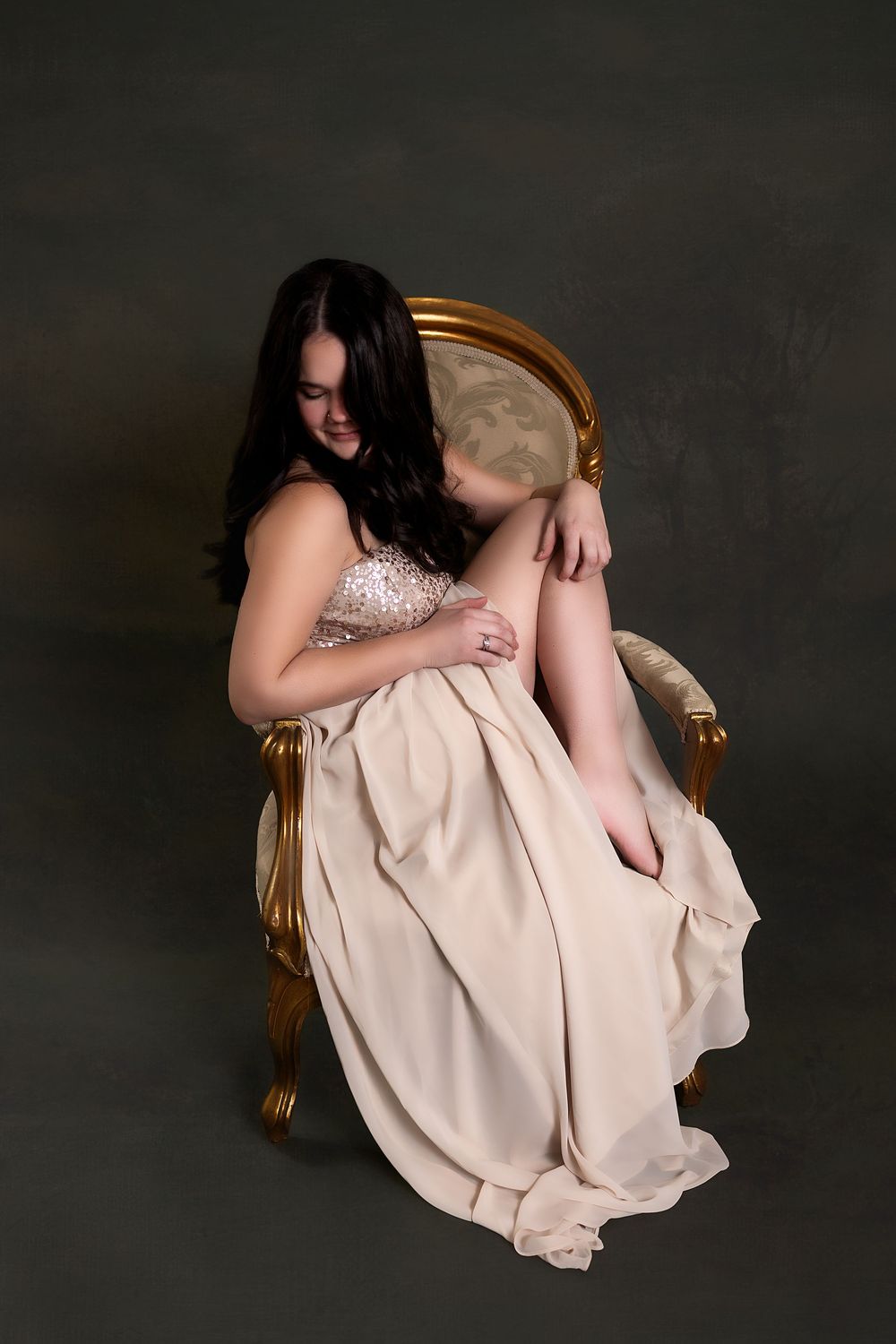 A woman with long dark hair sits in a gold chair, wearing a light-colored dress and gazing intently at the camera.