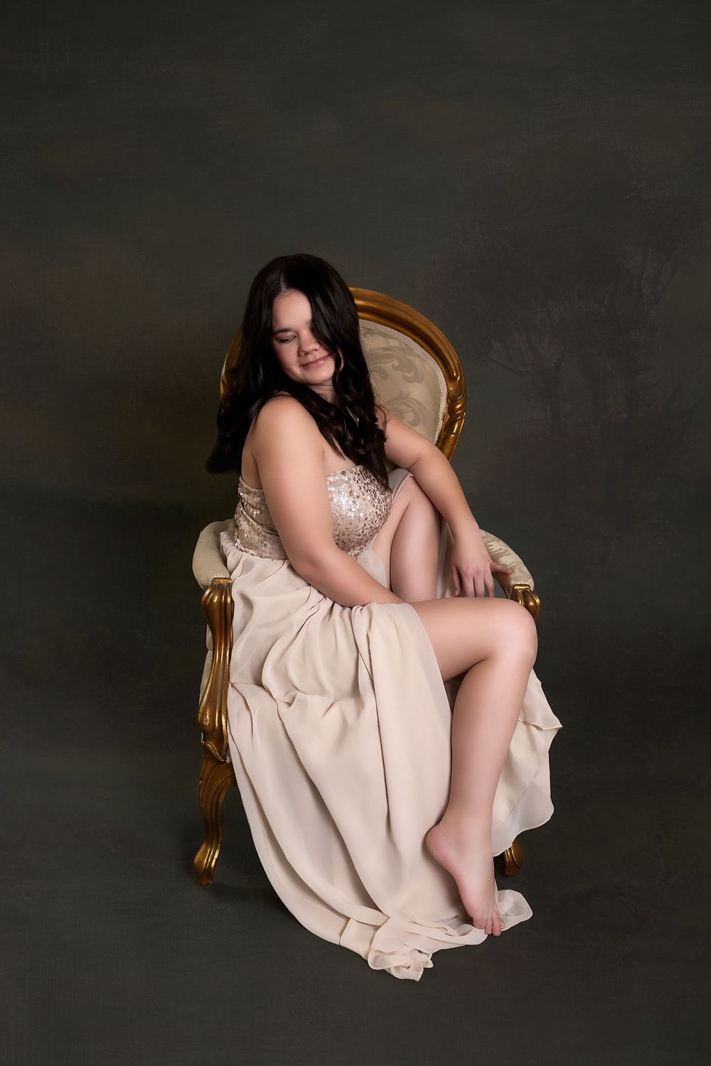 A woman in a beige dress sits in a chair with a thoughtful expression.
