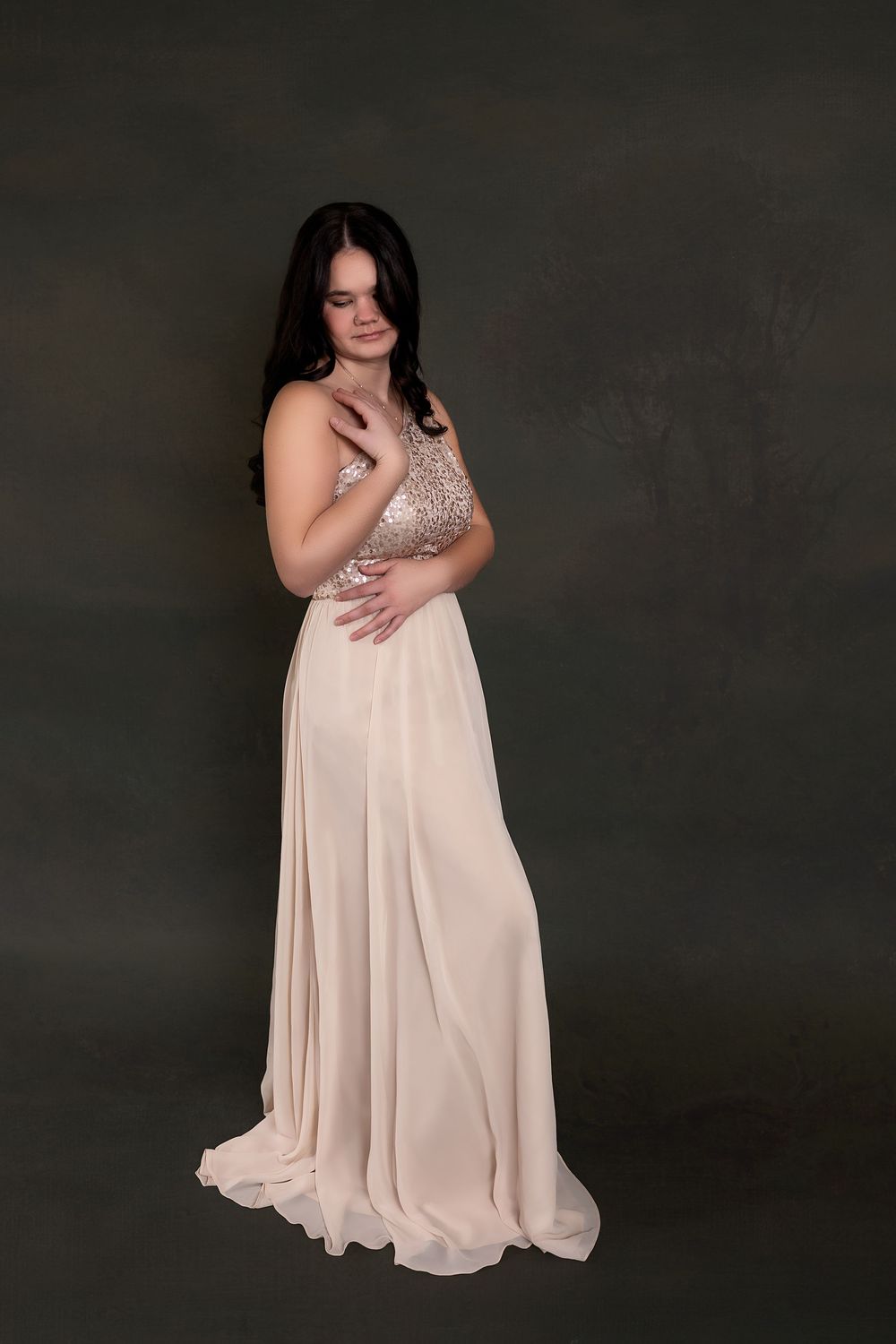 A woman in a long, flowing light-colored dress stands with her arms crossed in a dark studio setting.