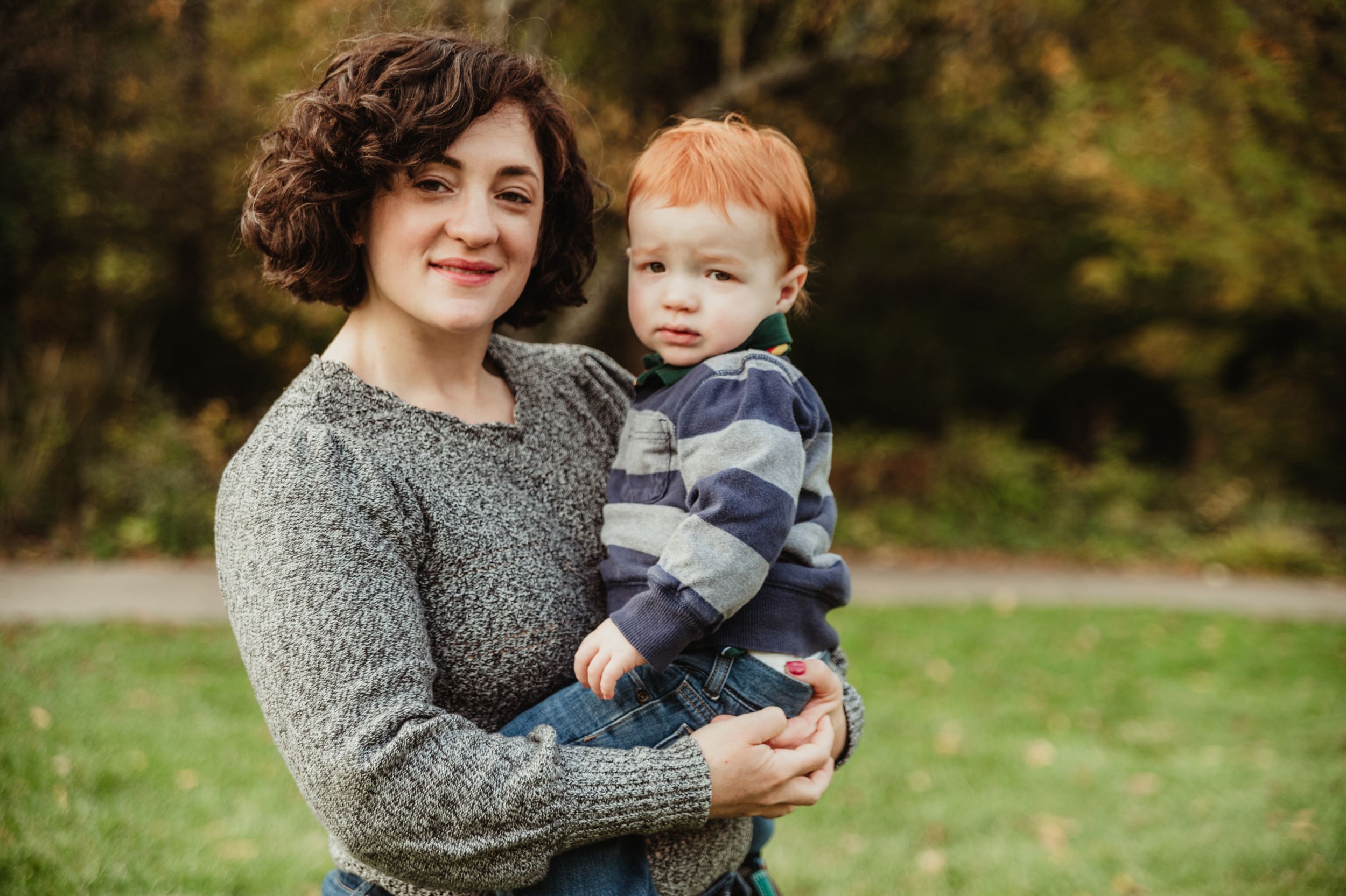 A woman in a gray sweater holds a young child wearing a striped shirt outdoors on a grassy field in autumn.