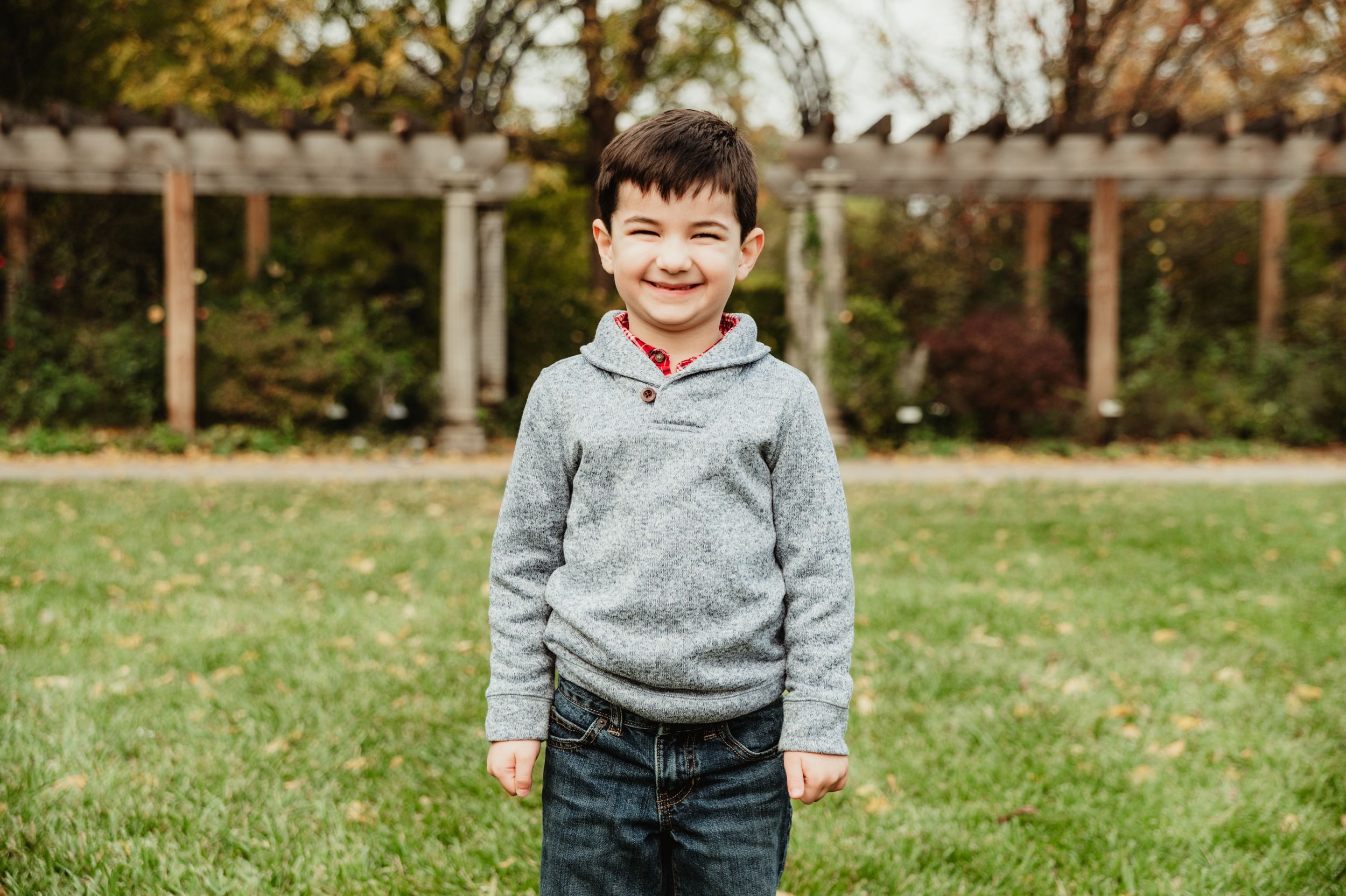 A cheerful figure stands on grass wearing a grey hoodie and jeans with a wooden fence in the background.