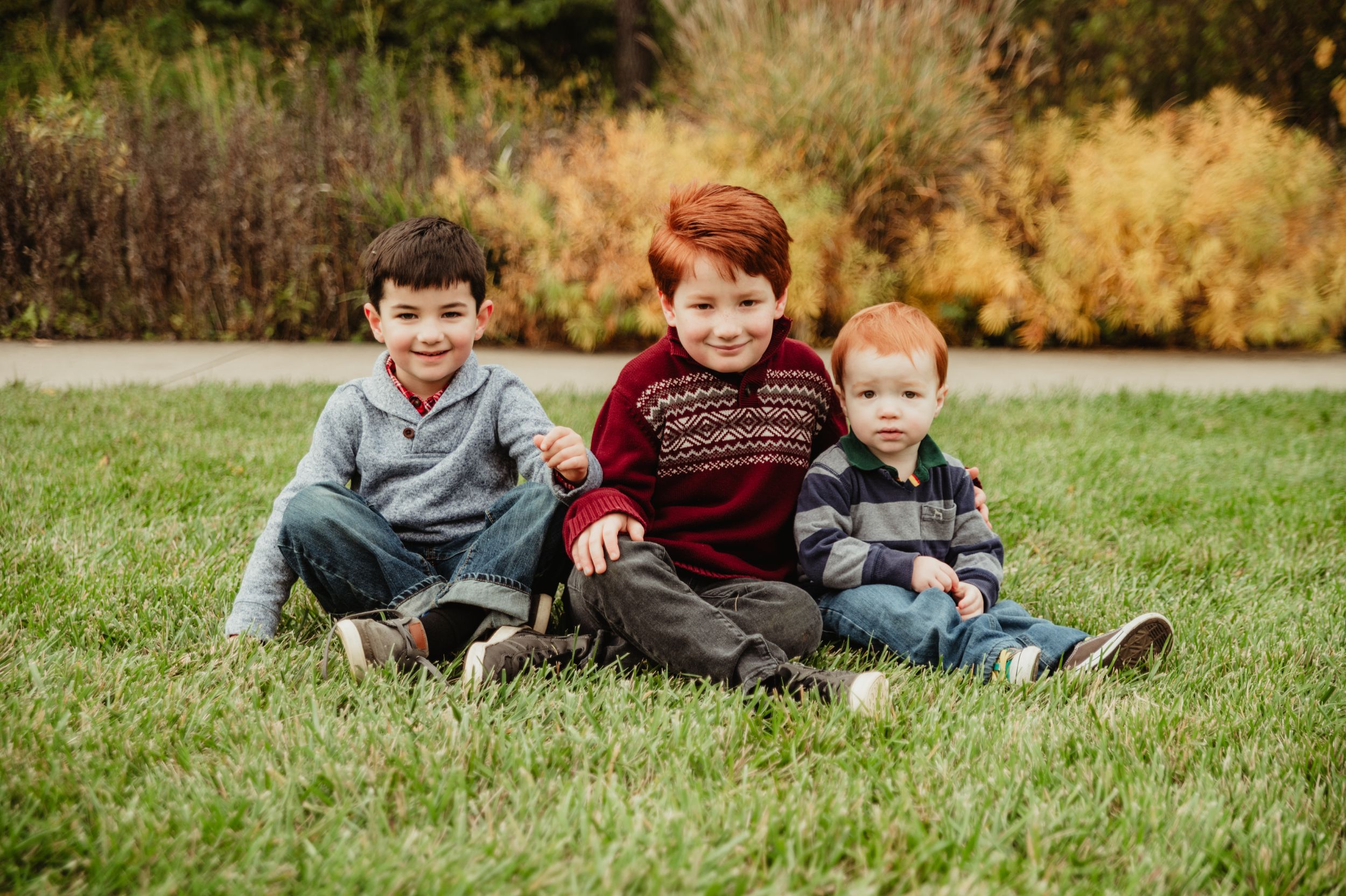 Three young children sit together on grass with golden autumn foliage in the background.