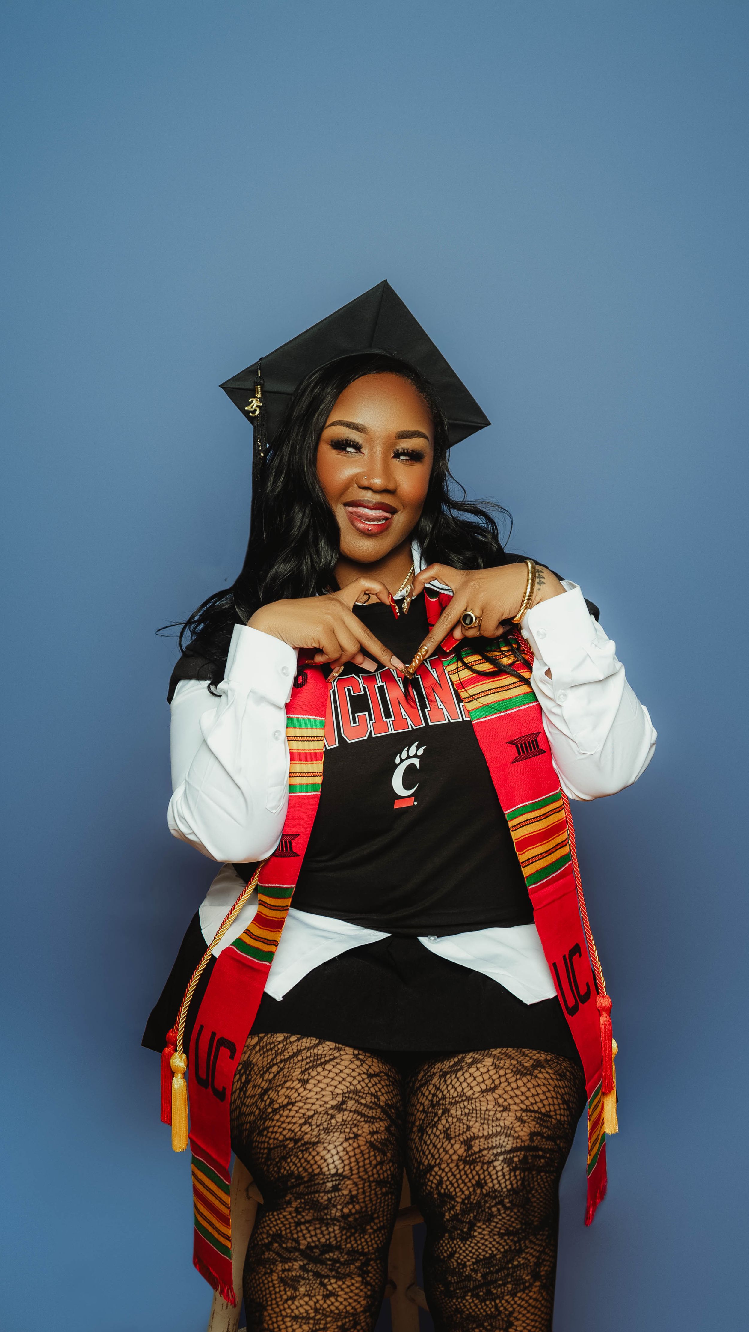 A graduation photoshoot featuring a graduate in cap and gown with colorful kente stole against a blue backdrop.