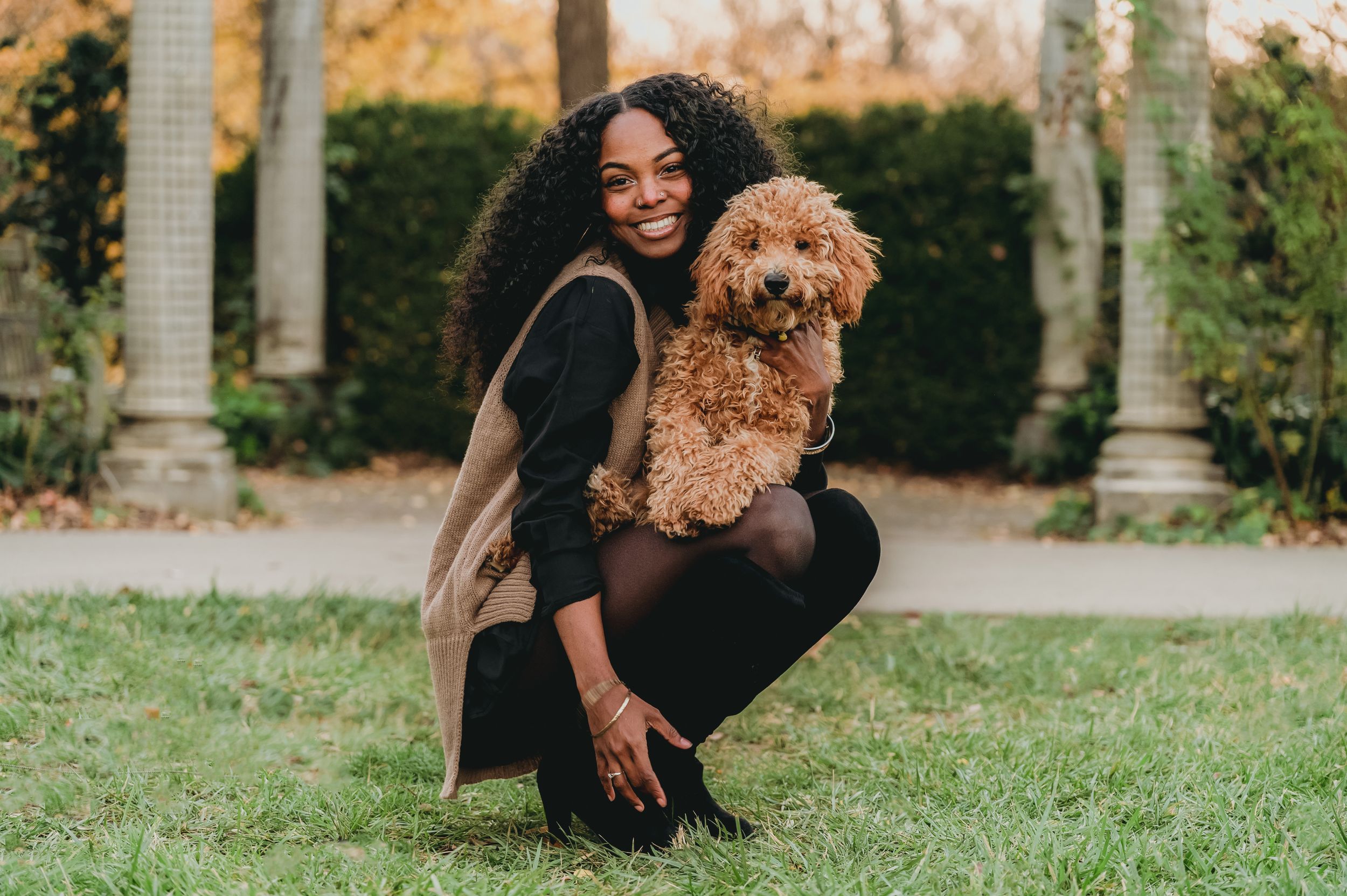 Someone in black clothing crouches on grass while holding a fluffy brown poodle mix dog in an outdoor garden setting.