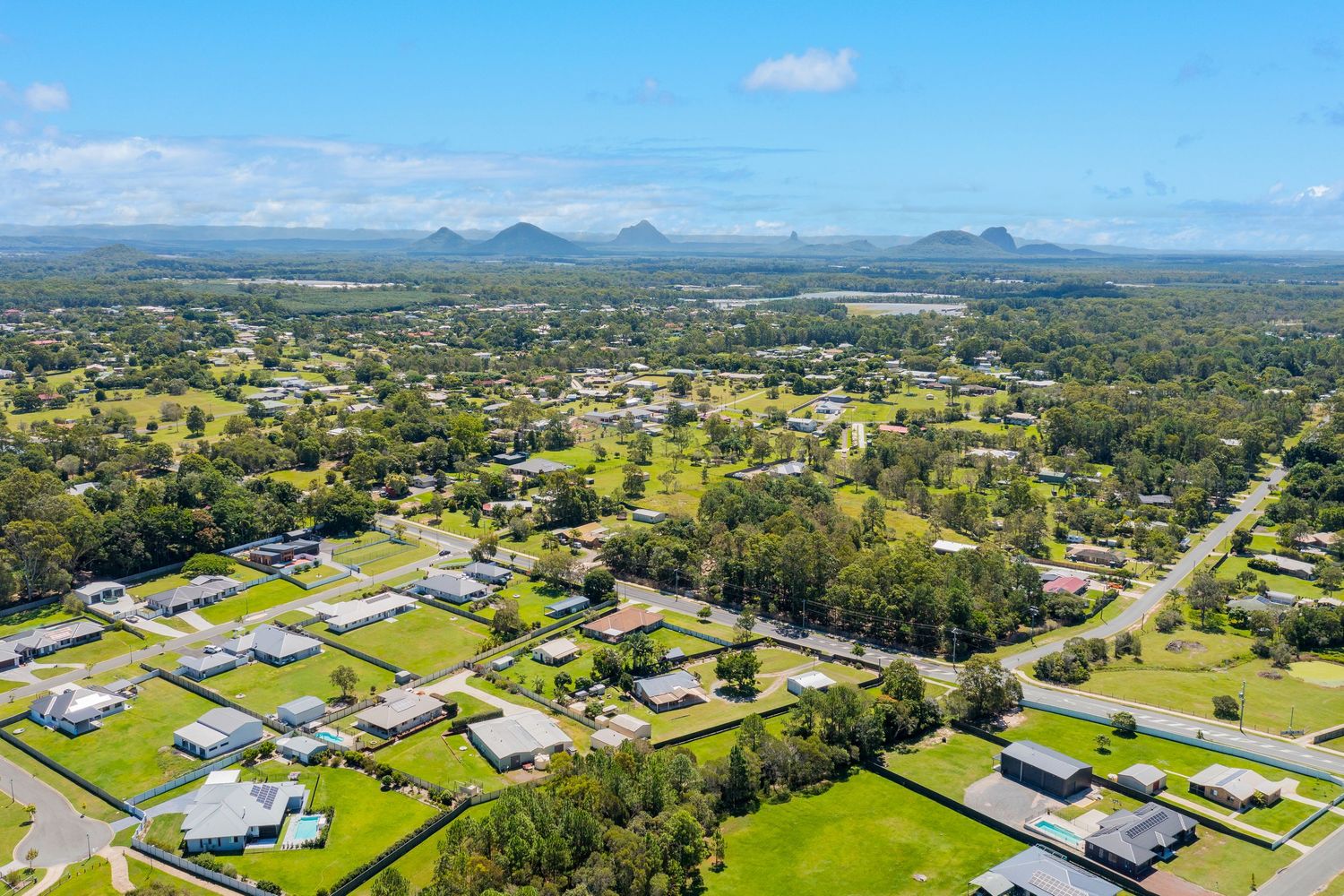 Aerial view of a suburban Queensland town with mountain peaks visible on the horizon under a clear blue sky.