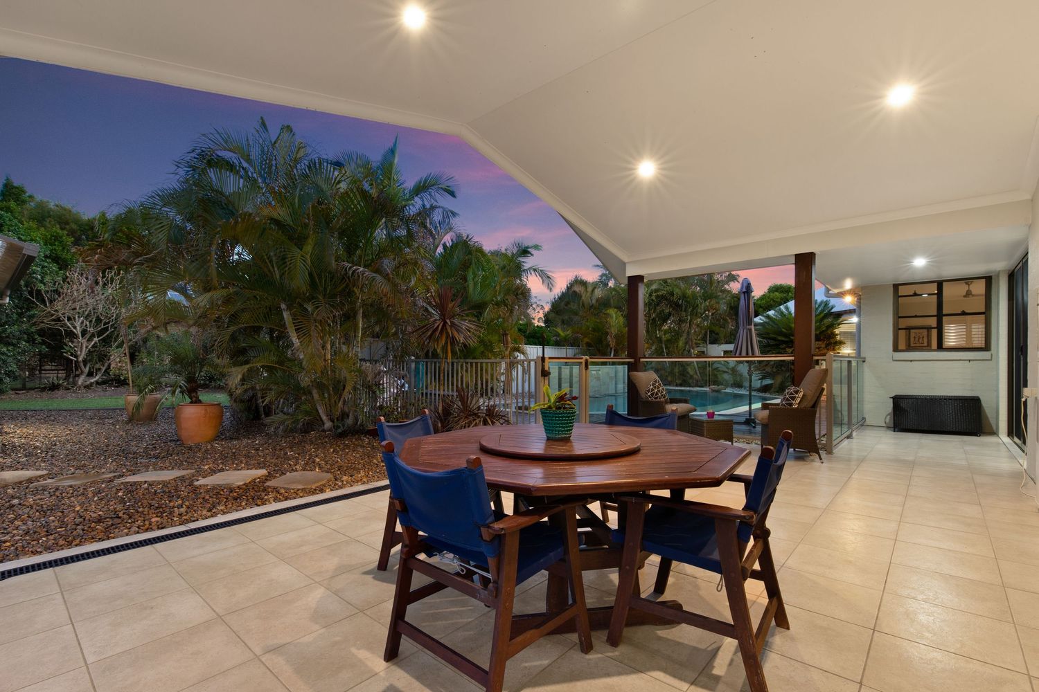Covered alfresco dining area with timber table overlooking tropical pool and garden.