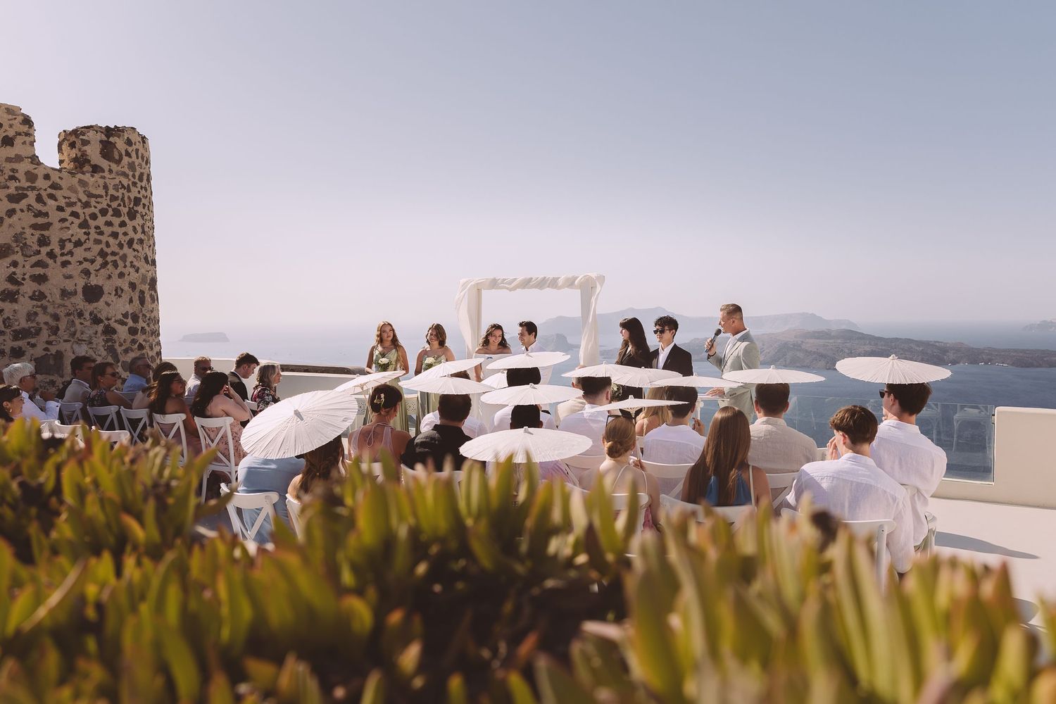 Wedding guests gather at a scenic coastal Santorini venue with stone walls and ocean views during a sunset celebration.