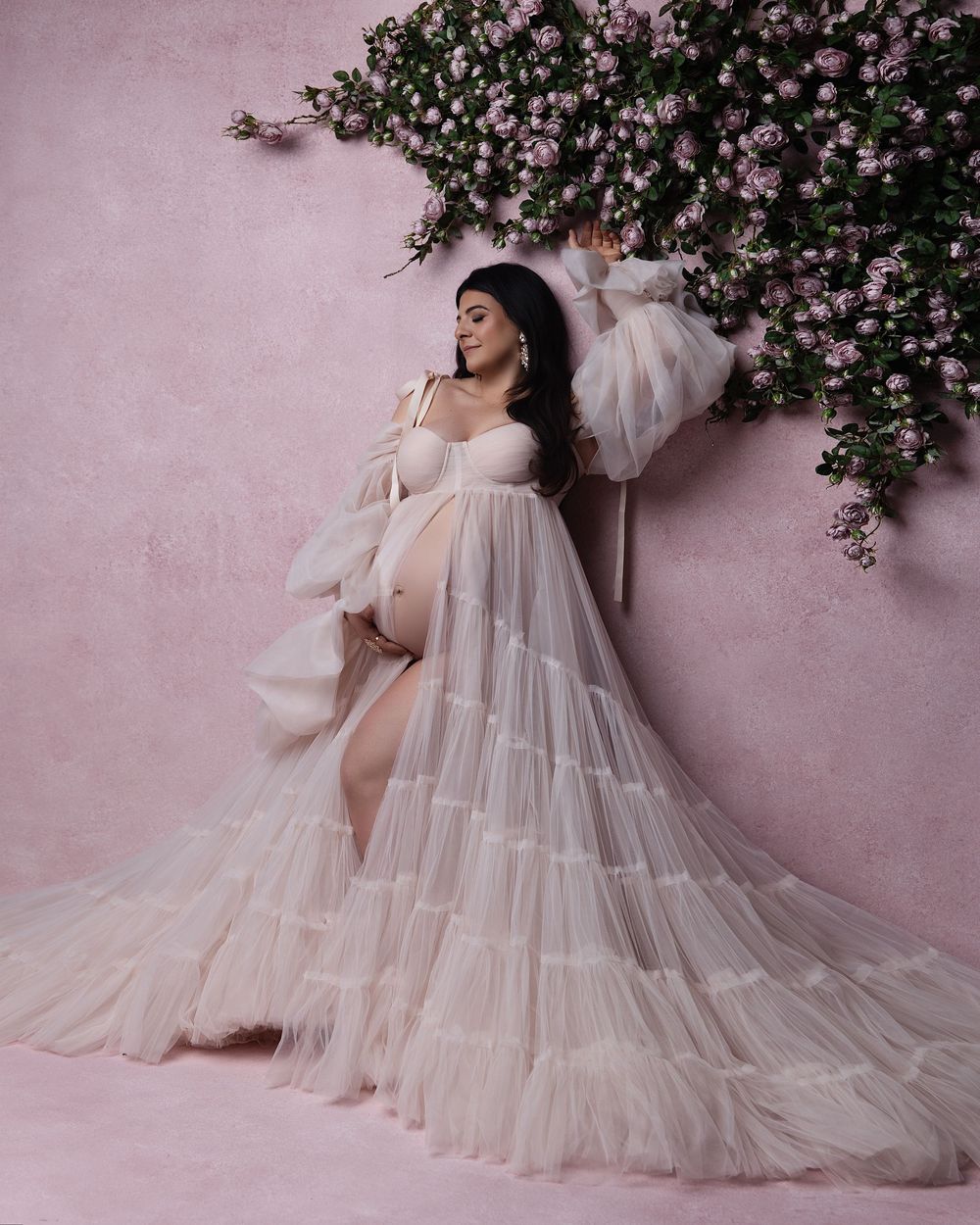 Pregnant woman in elegant white tulle maternity gown poses against pink wall with floral arrangement.