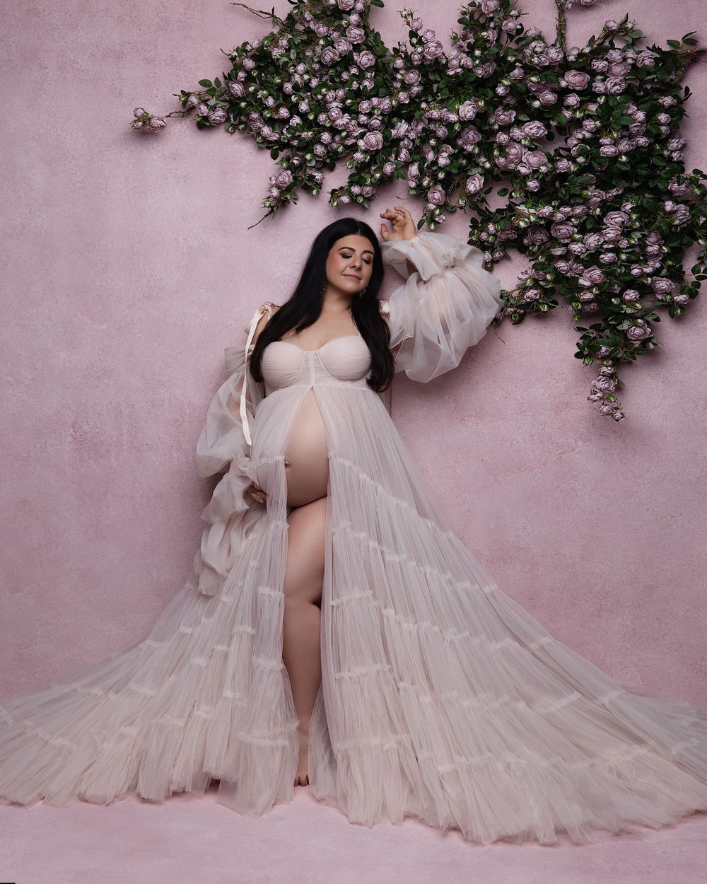 Pregnant woman in white tulle maternity gown poses against pink wall with floral backdrop for elegant maternity photo.