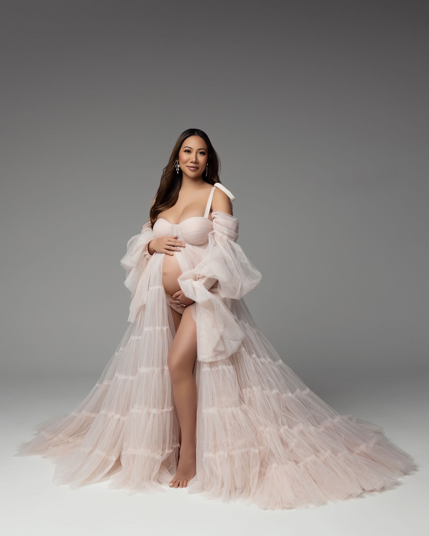 A graceful figure poses in a flowing blush-colored tulle gown against a gray background.