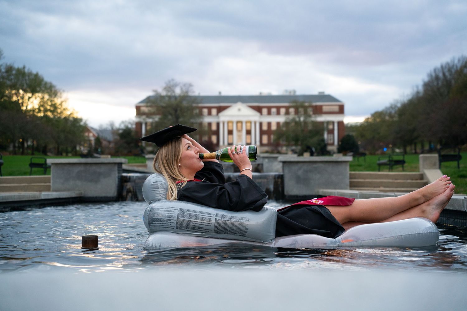 Graduate relaxing on a reflective surface near a stately building during sunset.