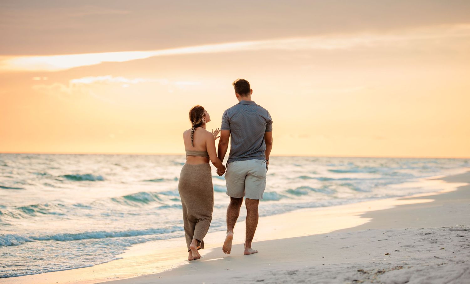 A couple walks hand in hand along a sandy beach at sunset as waves roll onto the shore.