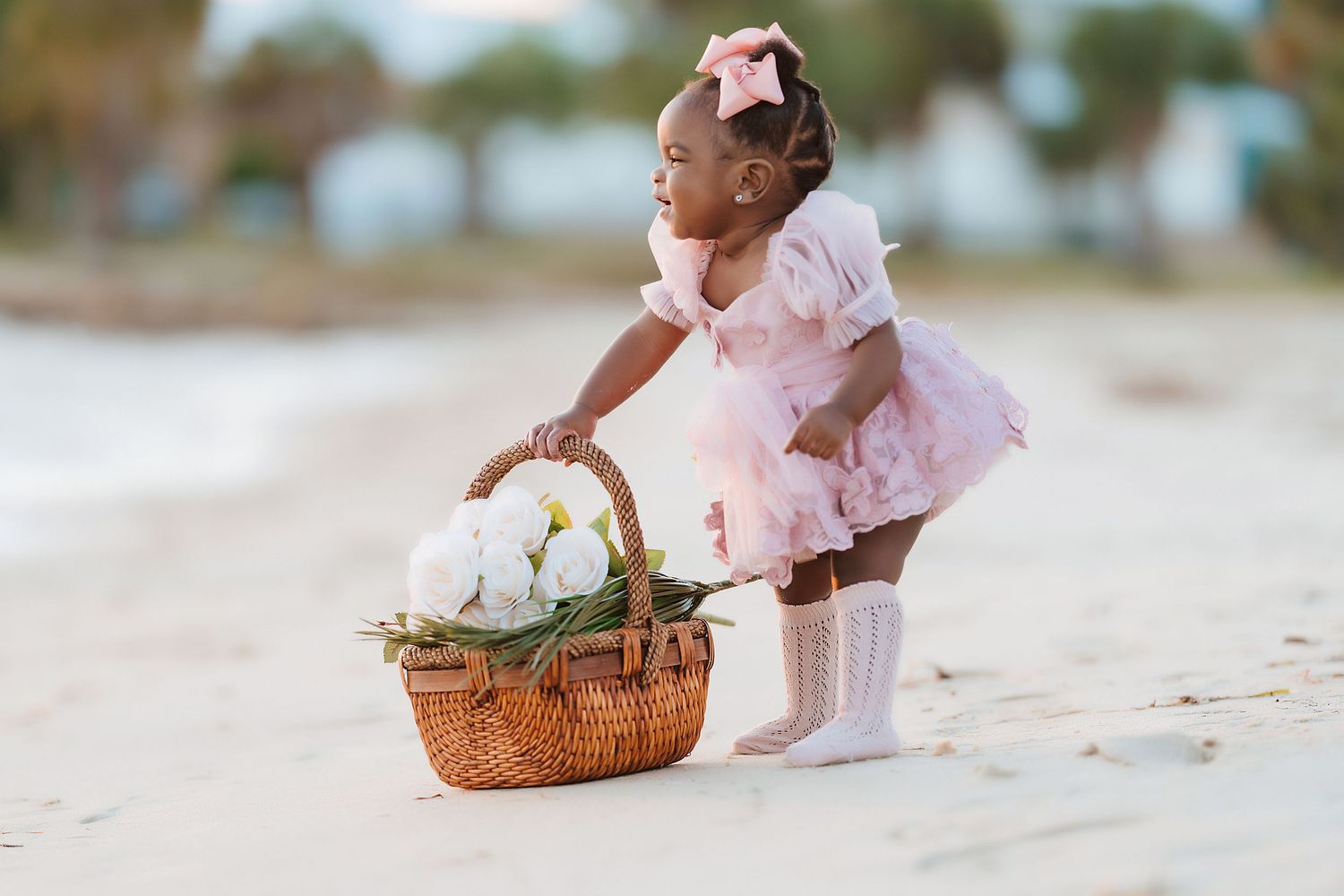 A child in a pink tutu dress bends down to pick up a wicker basket filled with white flowers on a sandy beach.