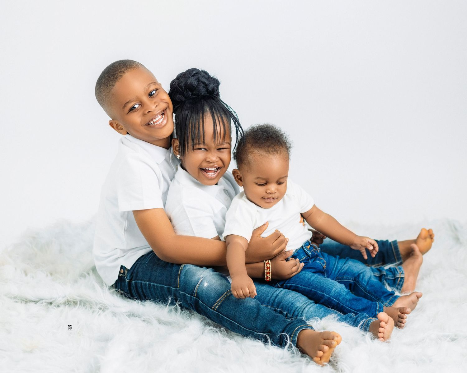 Three children in white shirts and blue jeans sit together laughing on a white backdrop during a studio photo session.