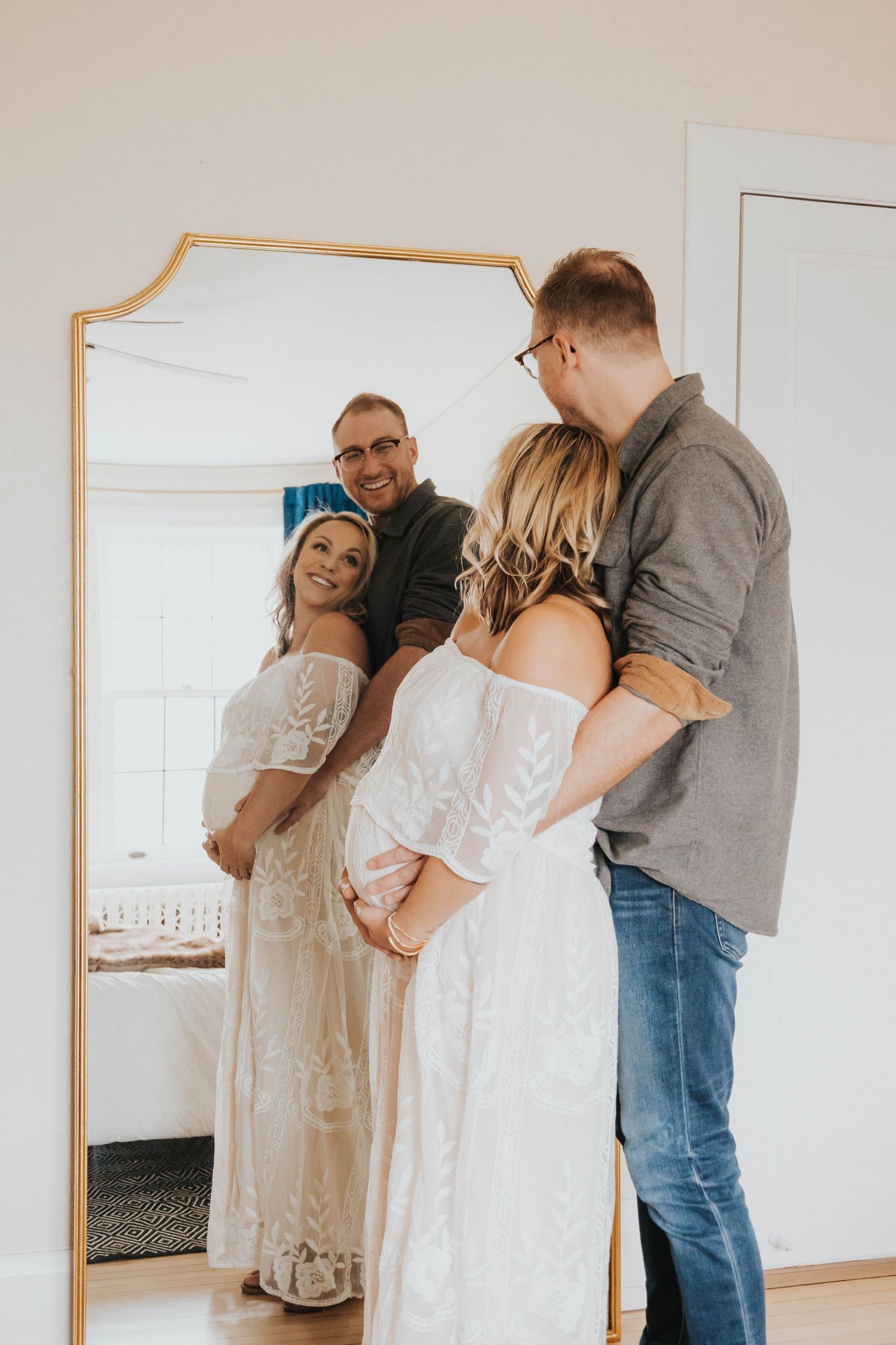 A couple in casual attire stands together posing in front of a gold-framed mirror in a bright interior space.