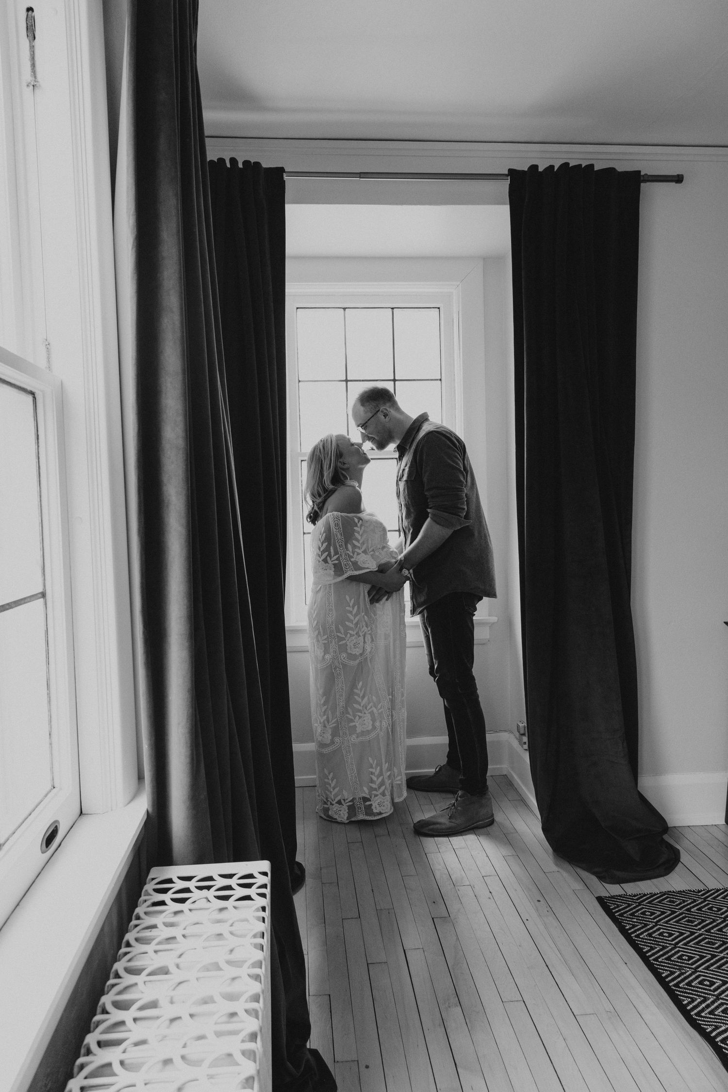 A romantic black and white photograph of a couple embracing by a window with long dark curtains.