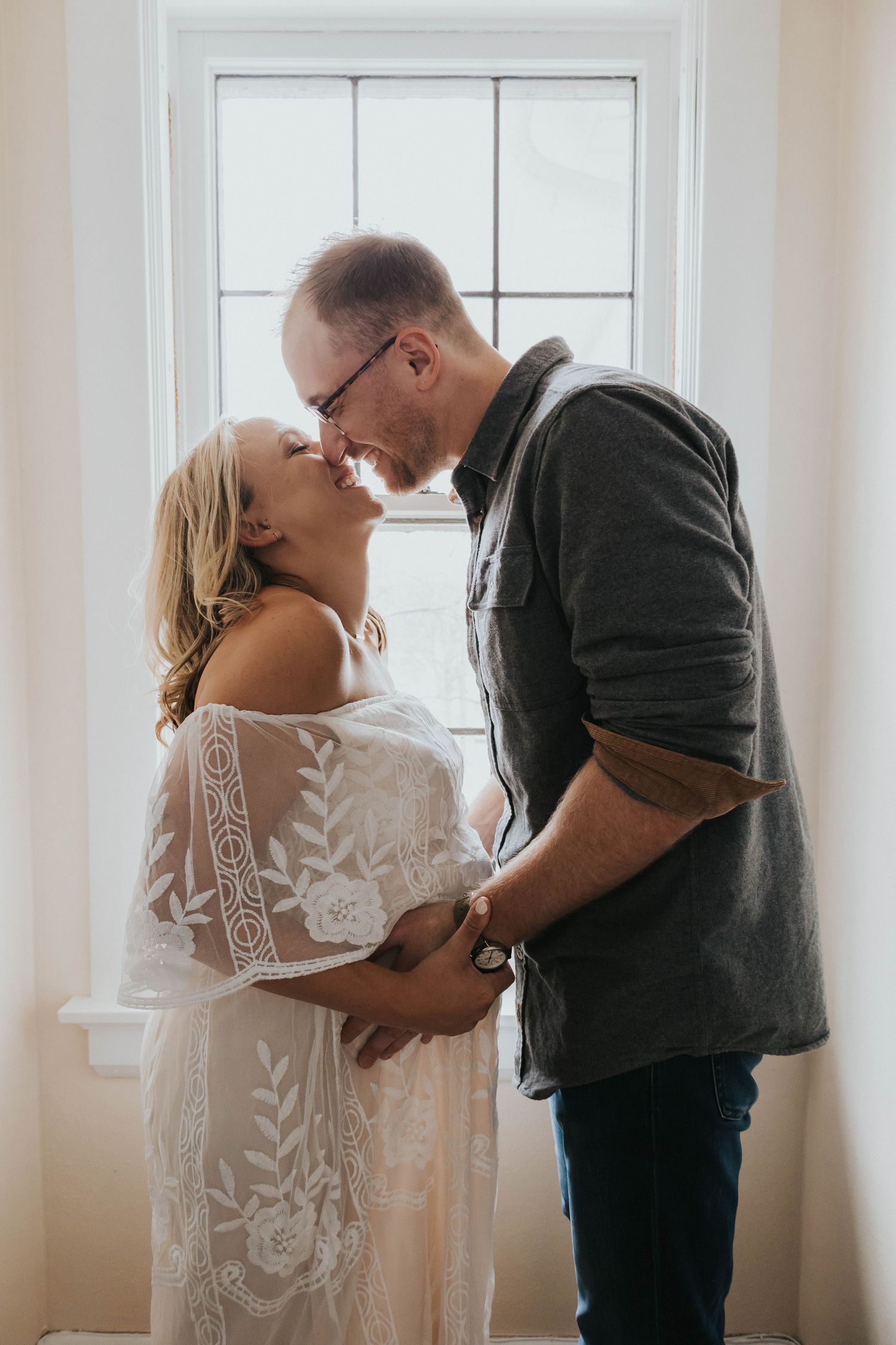 A tender moment shared between a couple by a window, with her in a white lace dress and him in a grey sweater.