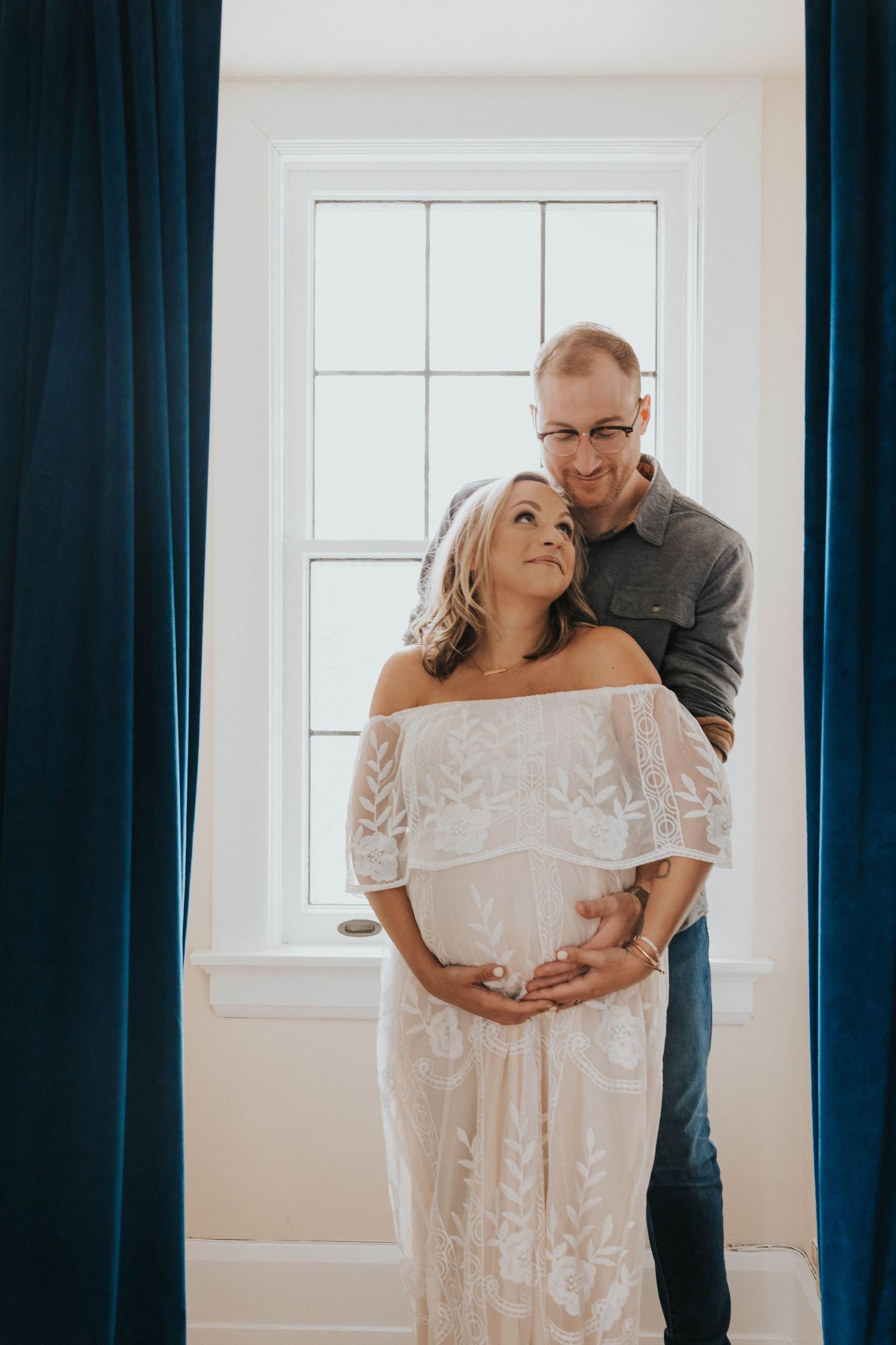 Intimate maternity photoshoot featuring a couple by a window with white curtains and off-shoulder lace dress.