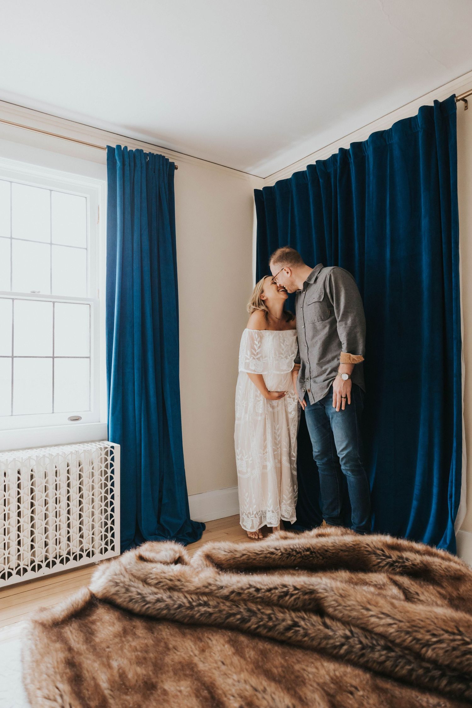 An intimate maternity photo of a couple standing by a window with navy blue curtains and a fur blanket.