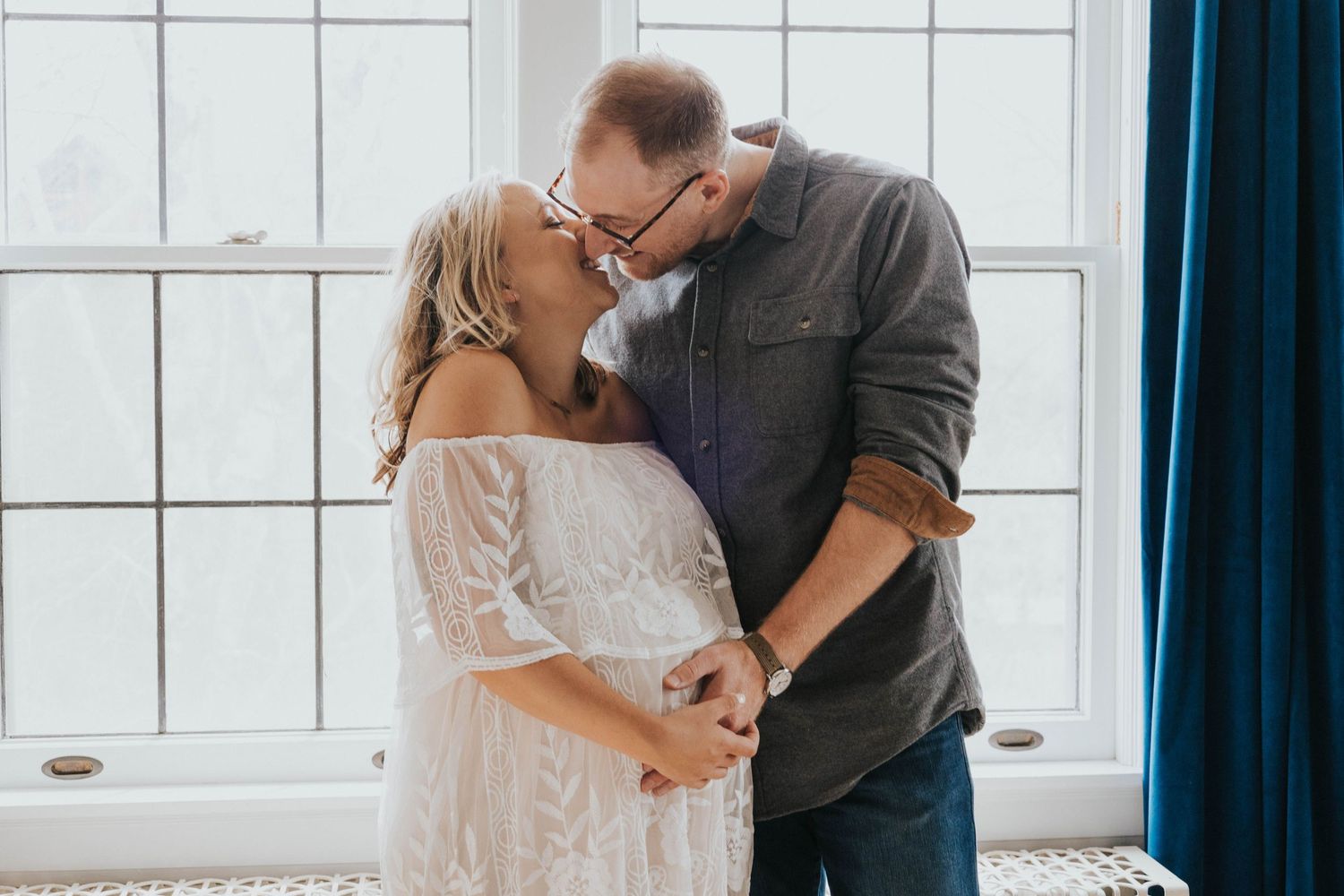 A couple in a white lace dress and grey shirt share an intimate moment by a bright window.