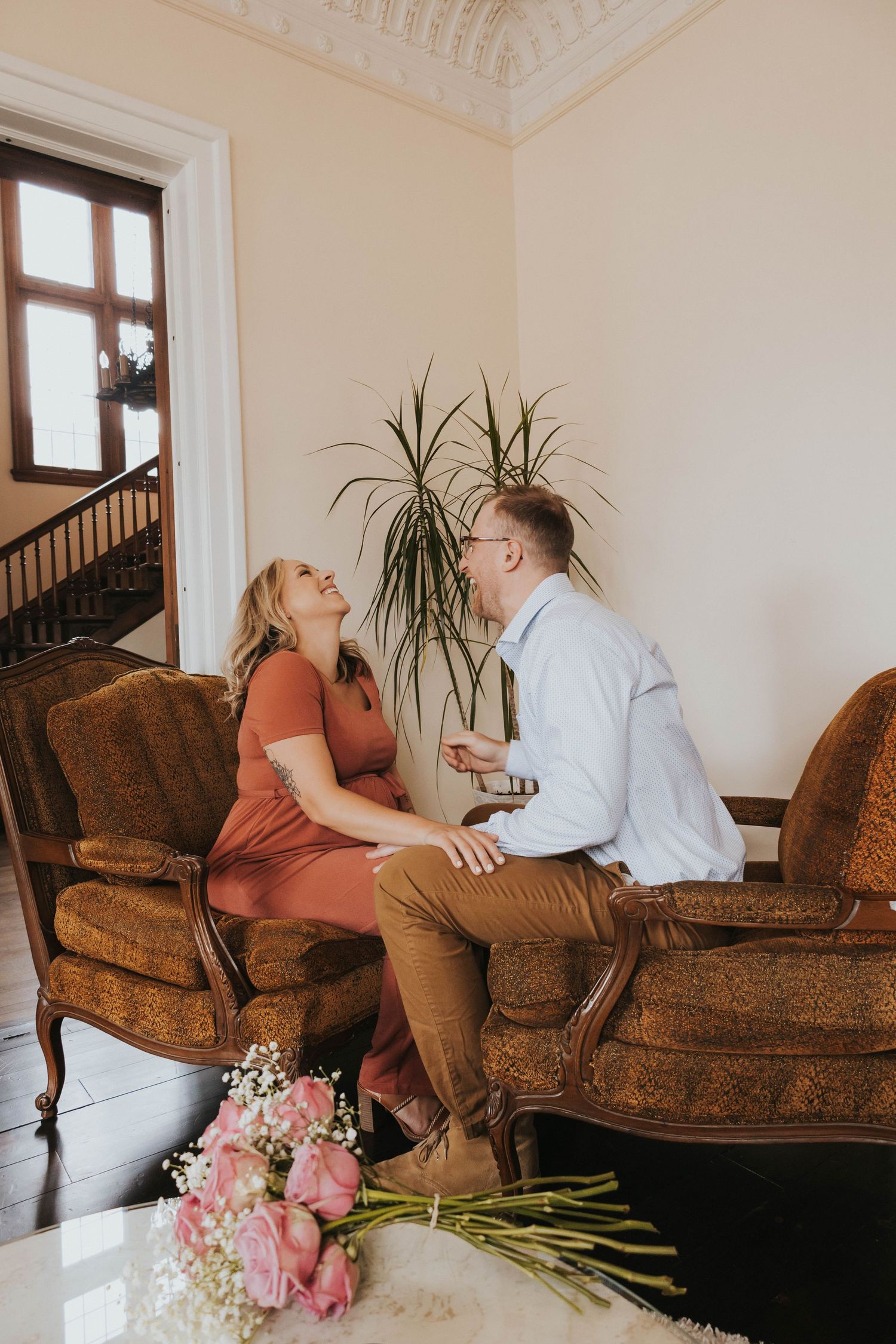 A romantic proposal scene with pink flowers on a vintage brown couch in an elegant room with ornate details.