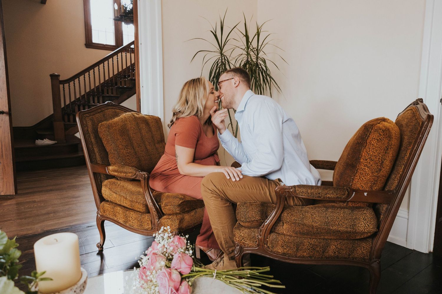 Couple sitting close together on vintage brown armchairs with pink flowers and candles nearby in an intimate setting.