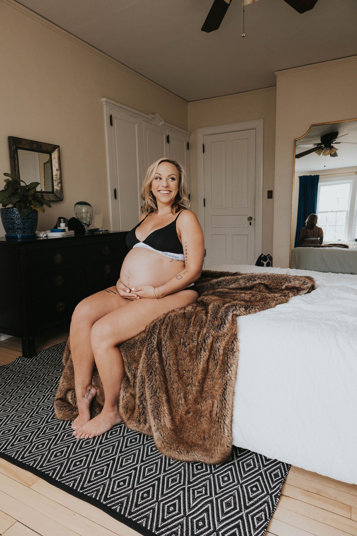 A pregnant woman in a black top sits on a bed with a brown fur blanket and geometric rug in a cozy bedroom setting.