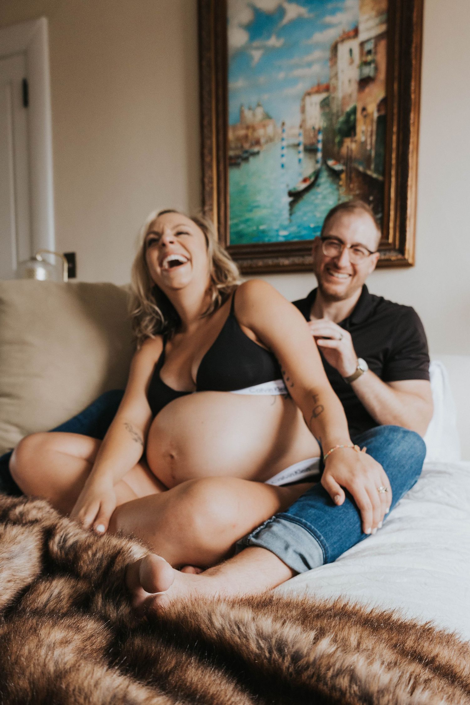 A joyful expecting couple shares a tender moment on a couch with a Venice canal painting in the background.