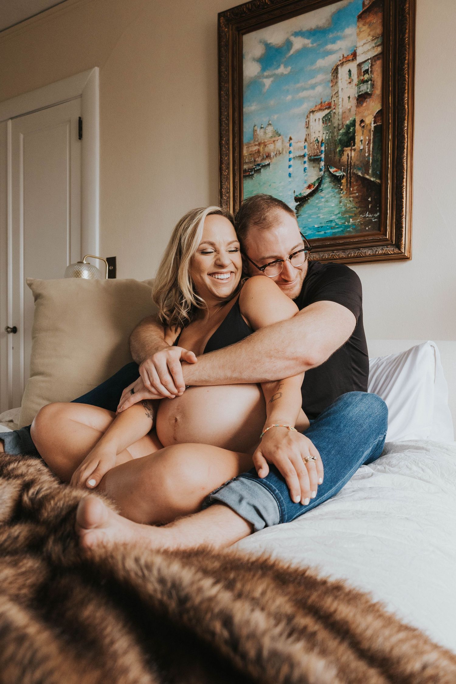 A couple cuddles on a bed with a brown fur blanket as they relax in a bedroom featuring a Venice canal painting.