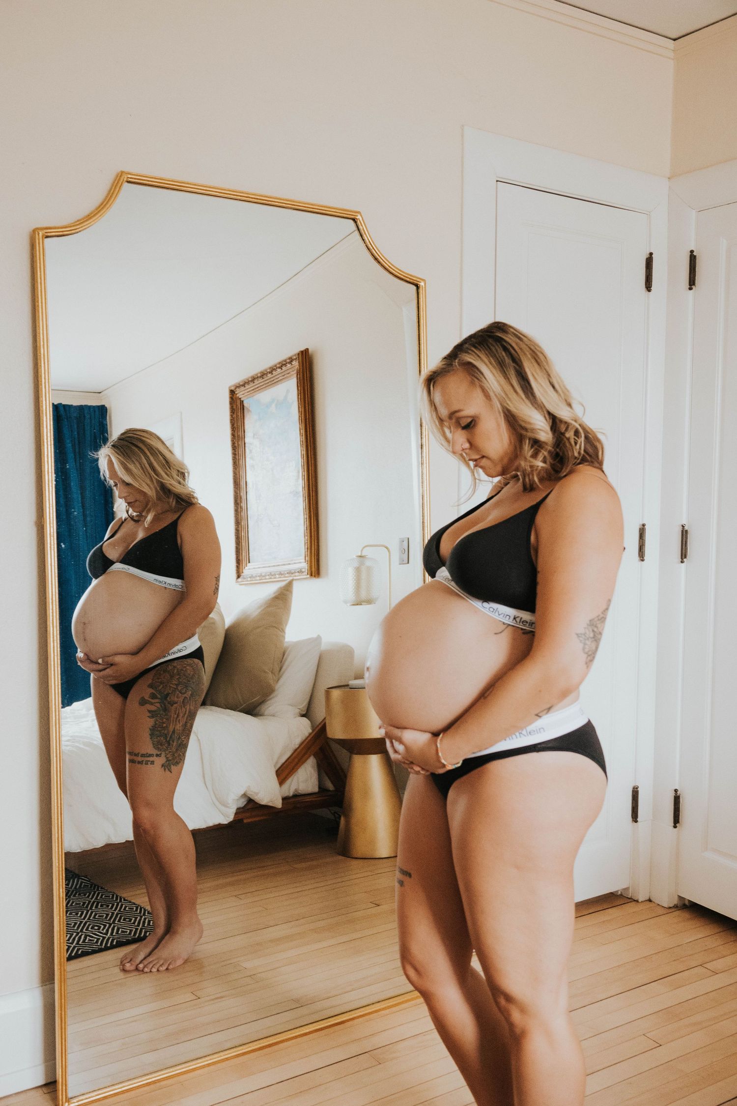 Pregnant woman stands sideways admiring her baby bump reflection in a full-length mirror with gold frame.