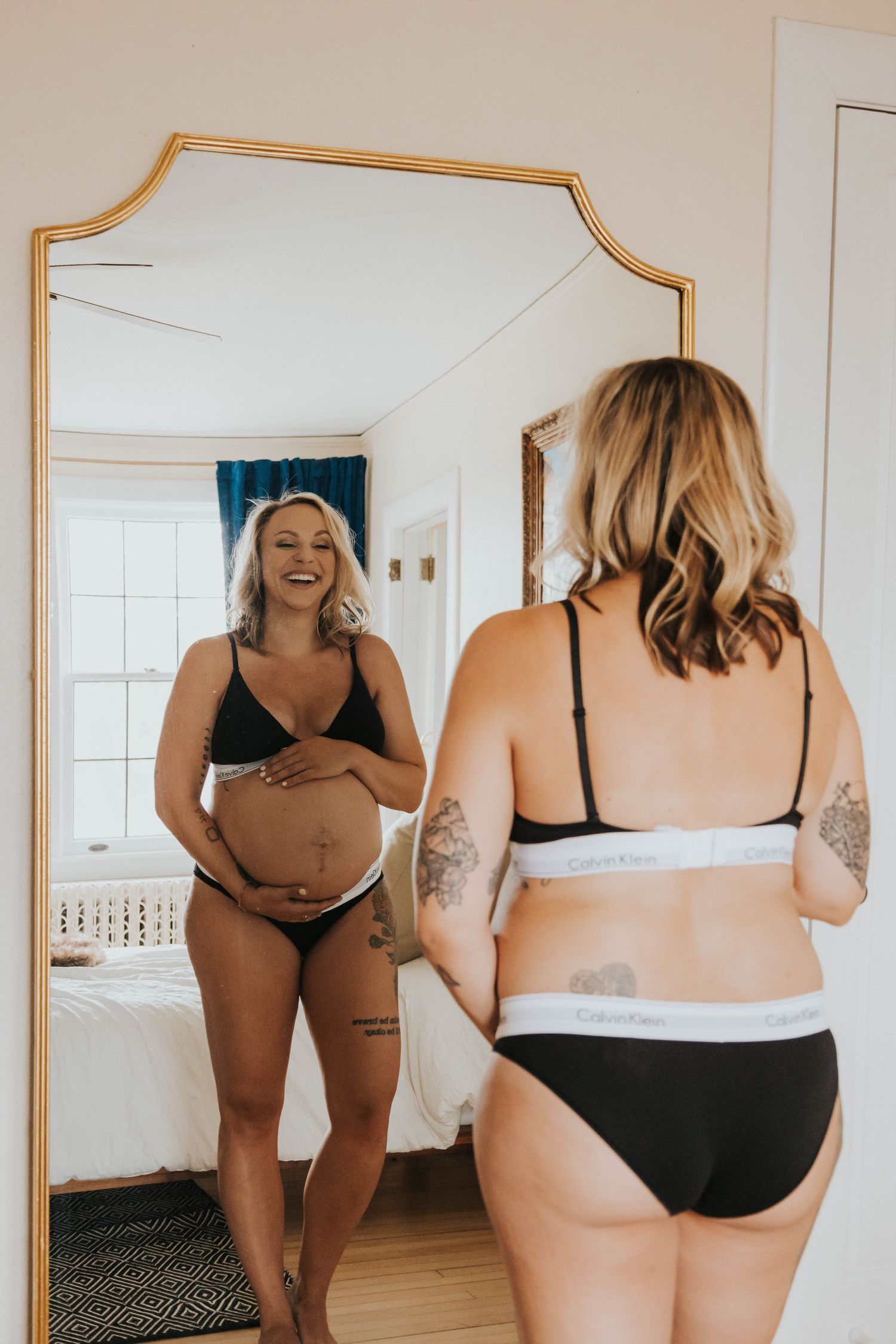 A pregnant woman in black lingerie looks at her reflection in a mirror while touching her belly.