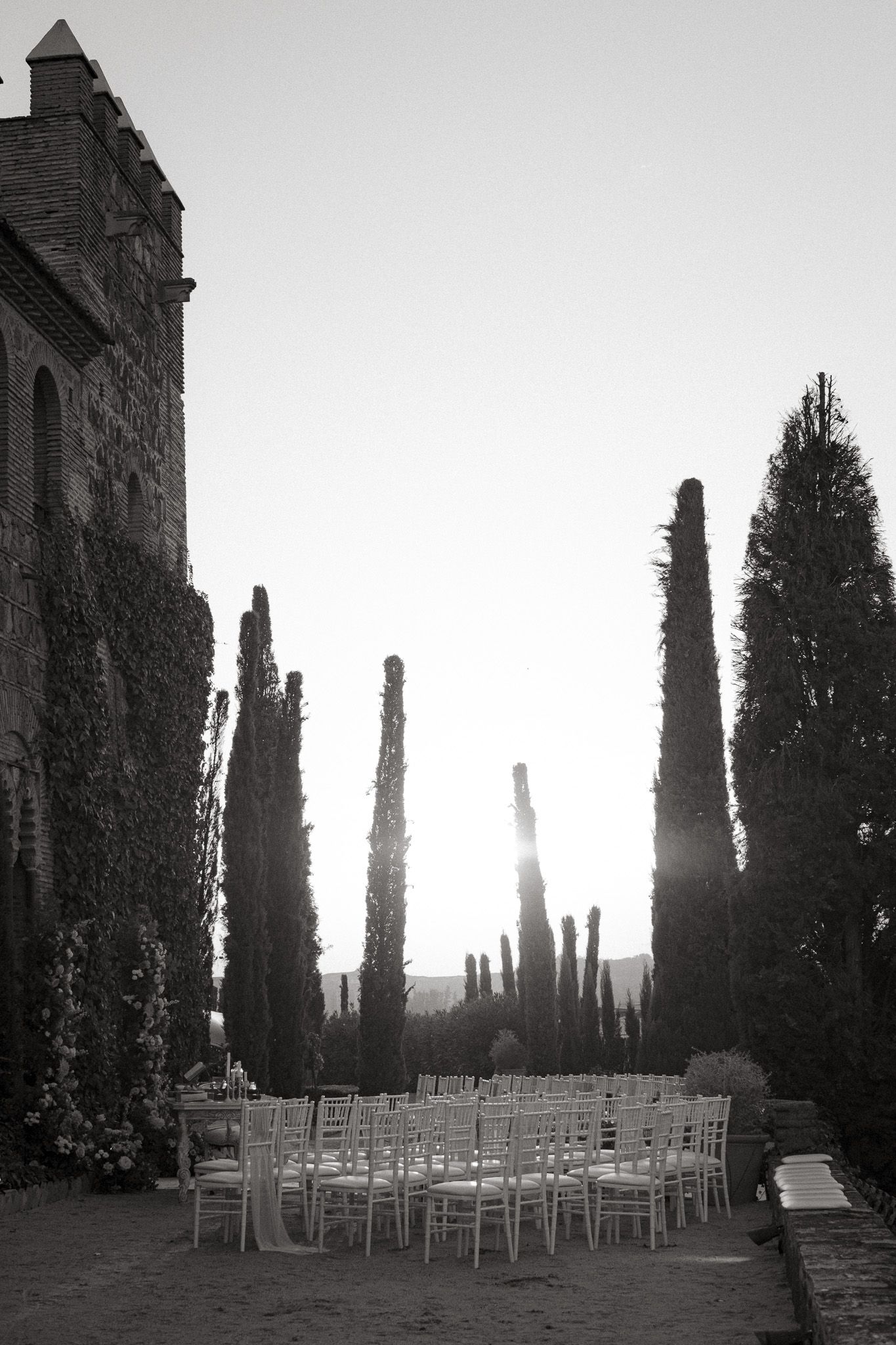 White chairs arranged for an outdoor wedding ceremony against tall cypress trees and historic building at sunset.