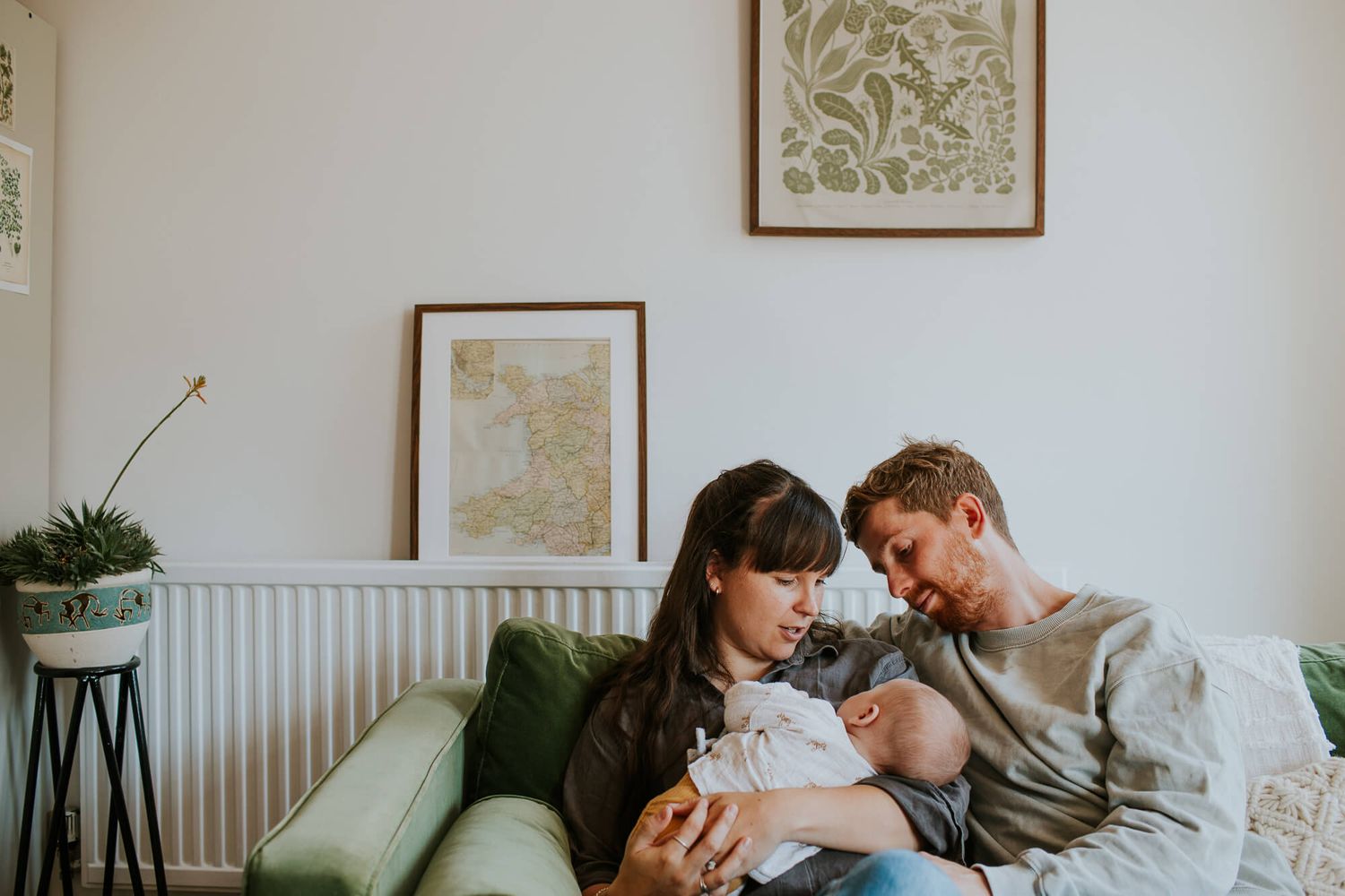 Couple relaxing on green sofa with baby at home, framed artwork and plant on stand in background.