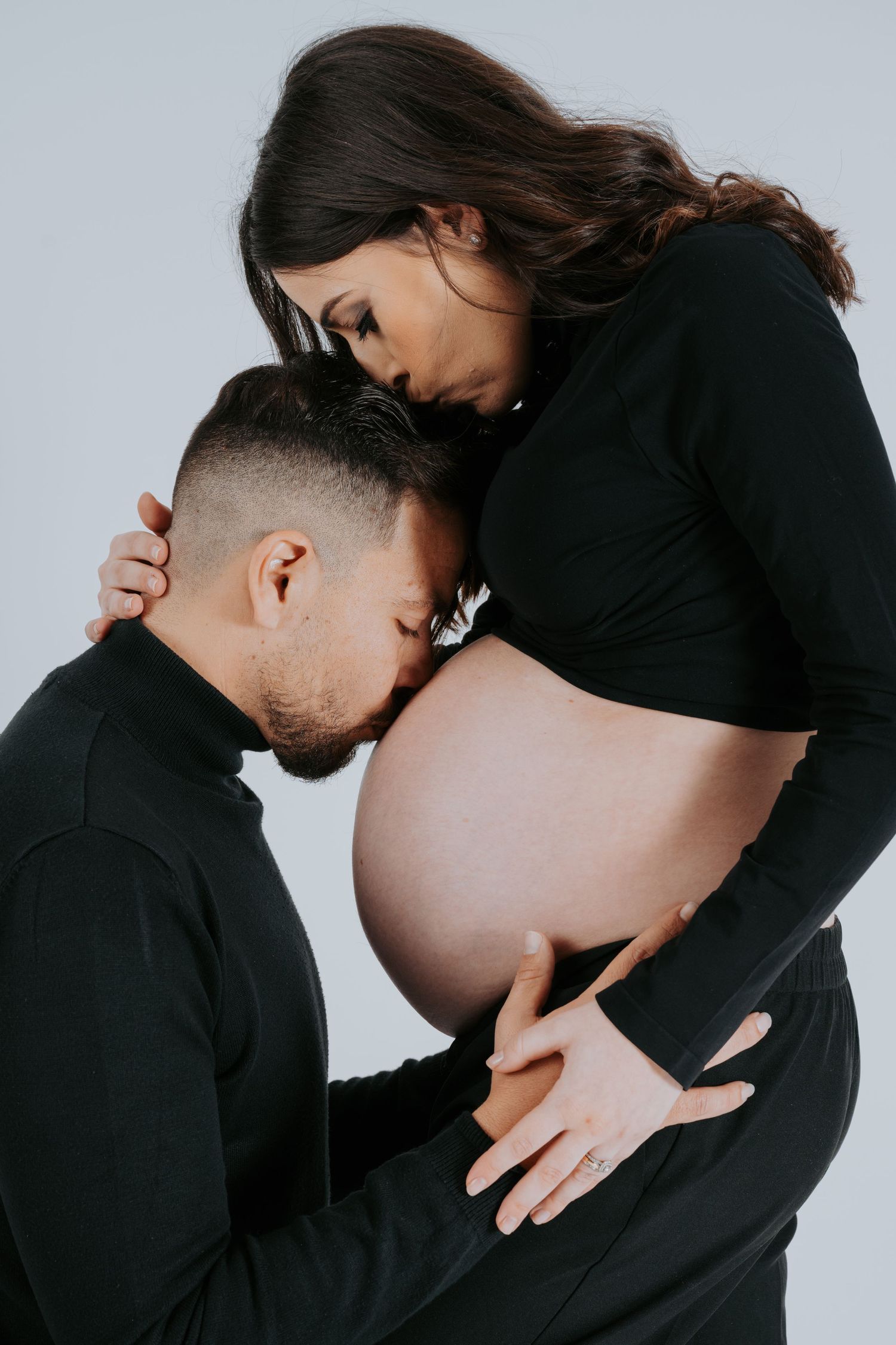 Intimate maternity photoshoot featuring couple in black outfits against neutral background.