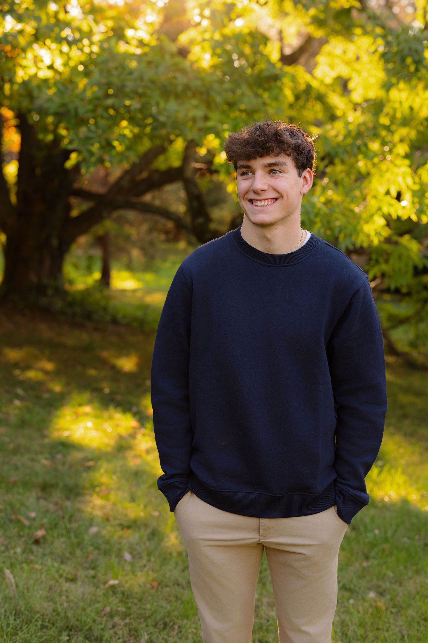Someone in a navy blue sweater and khaki pants smiles while standing in front of fall foliage in an outdoor setting.