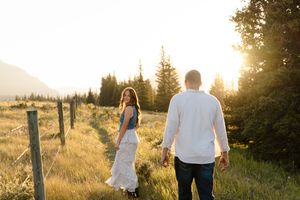 Romantic couple walks through golden meadow at sunset while holding hands in scenic mountain landscape.