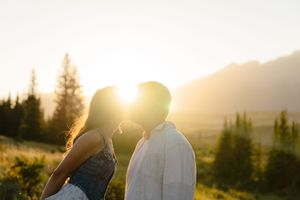 A couple embraces in a golden sunset with mountains and evergreen trees silhouetted in the background.