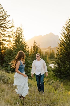 A couple walks through a mountain meadow at sunset with evergreen trees and peaks silhouetted in the golden light.