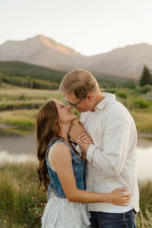 A romantic couple embraces and kisses in a scenic mountain landscape with a lake and tall grass at sunset.