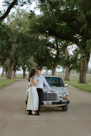 A couple poses romantically by a vintage Mercedes-Benz on a tree-lined road during a sunset photoshoot.